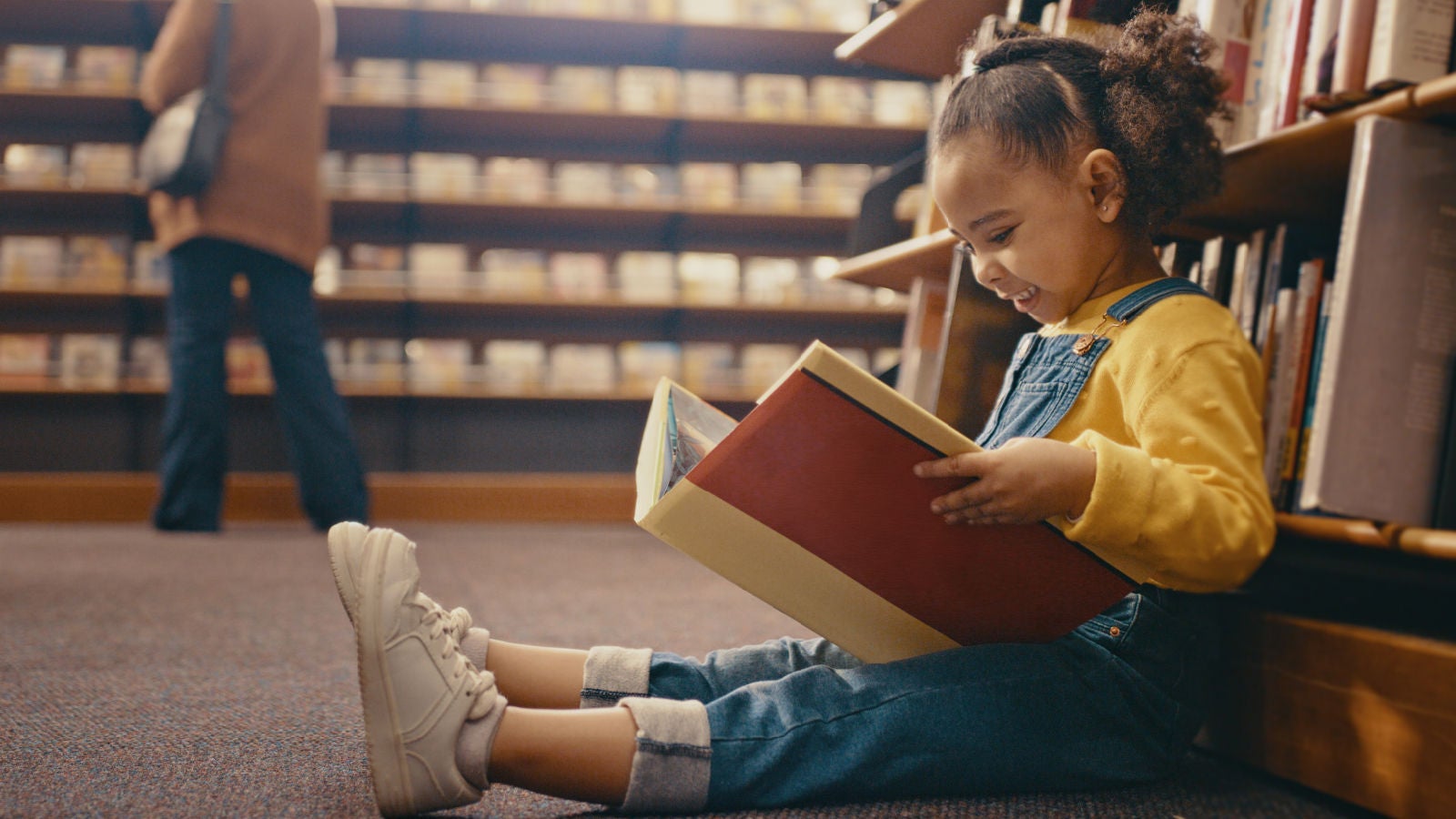 A young girl in a yellow sweater and denim overalls sitting on the floor of a library, smiling as she reads a large book.