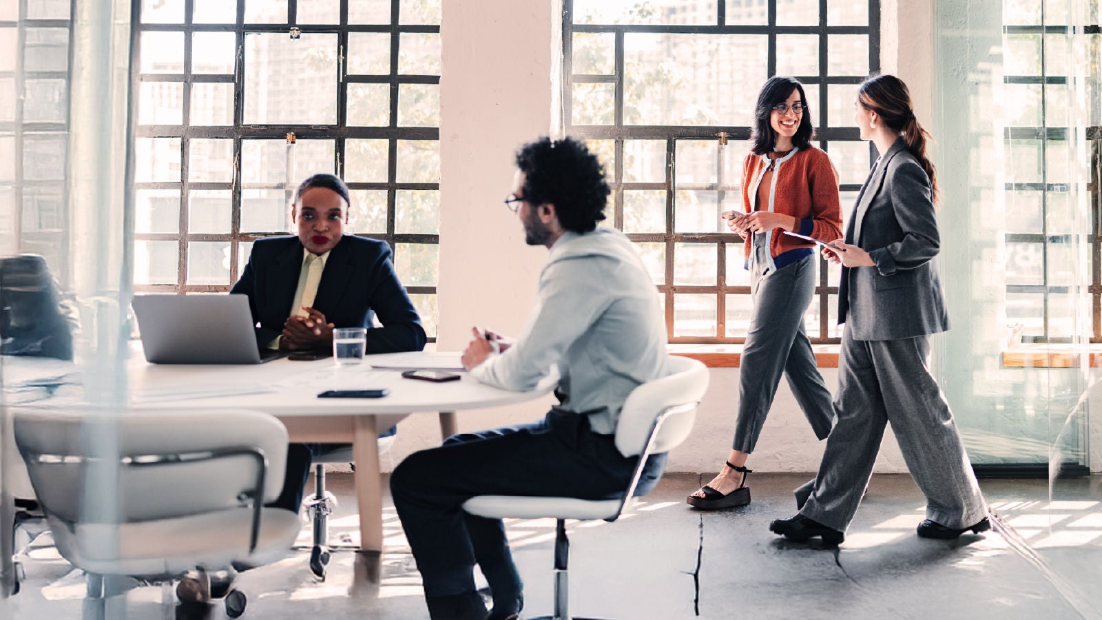 A group of colleagues working in a bright office, with two women walking and talking near large grid windows while others sit at a meeting table.