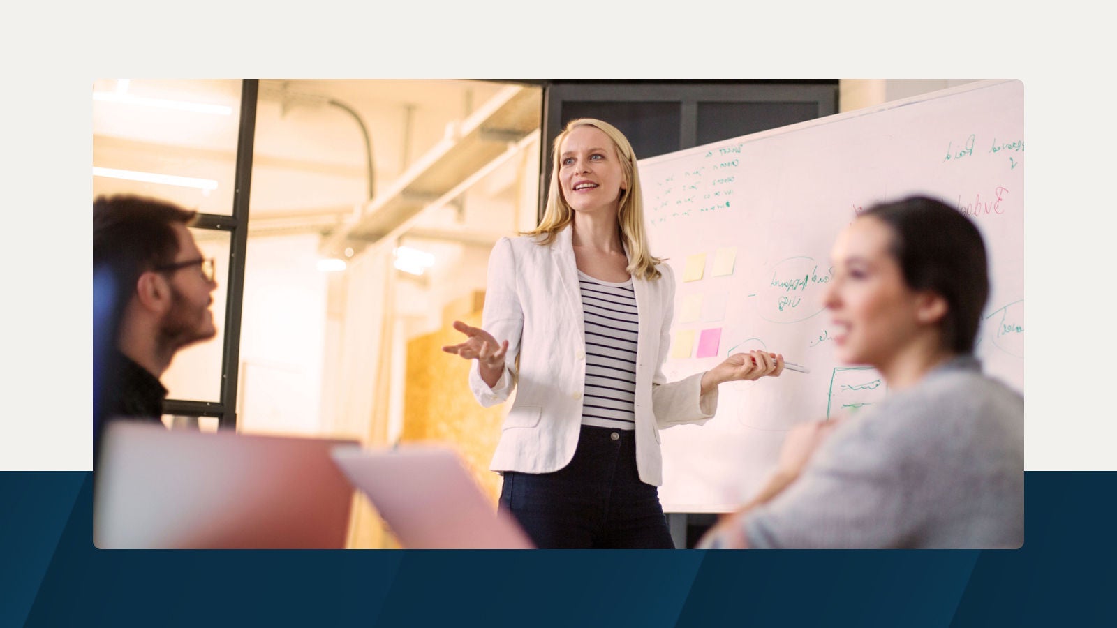 A woman in a white blazer leads a discussion in front of a whiteboard filled with handwritten notes and sticky notes, while two colleagues listen and engage during a collaborative office session.   2/2