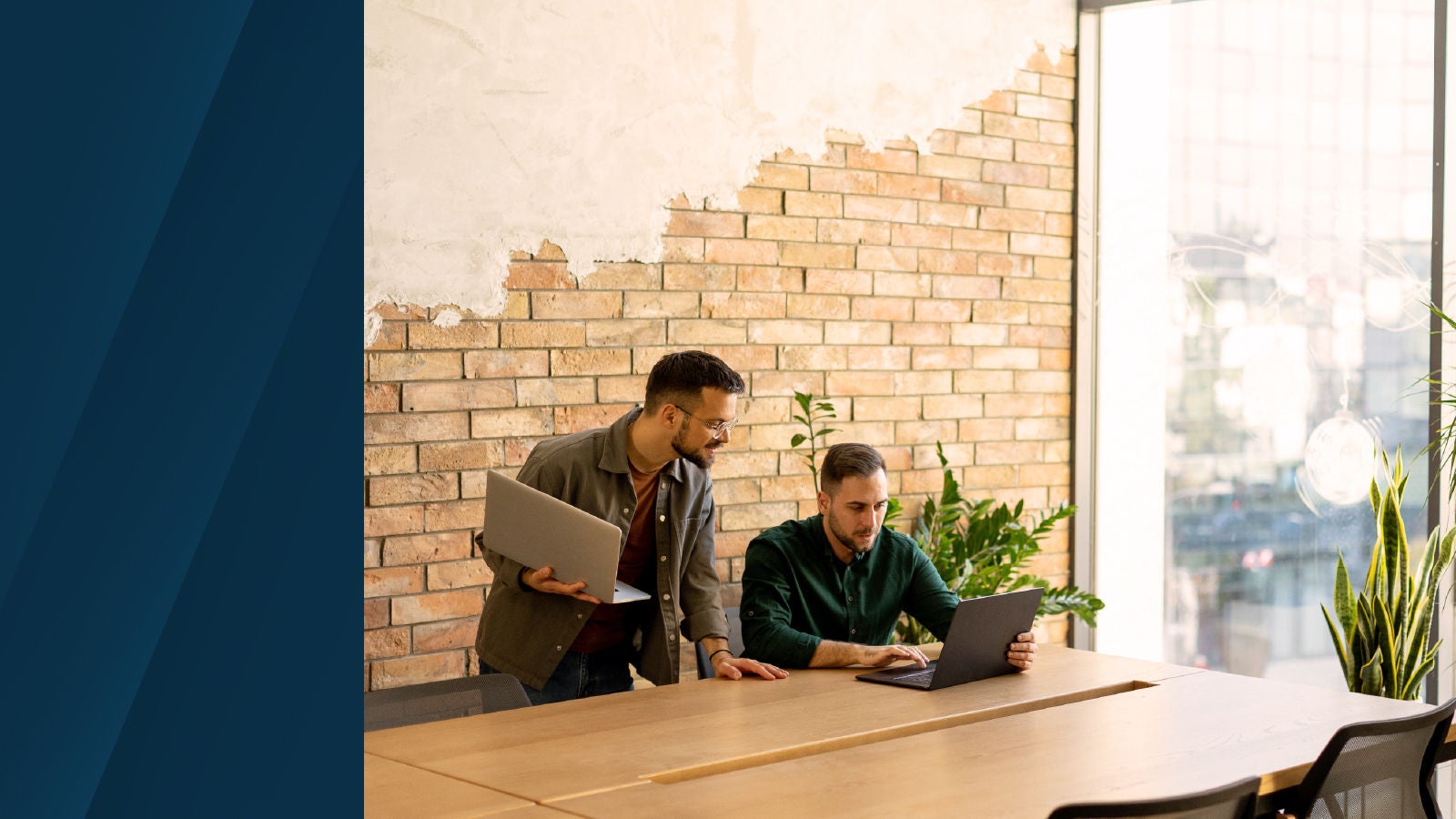 Two professionals working together in a bright, modern office with exposed brick walls and large windows, both focused on a laptop screen.