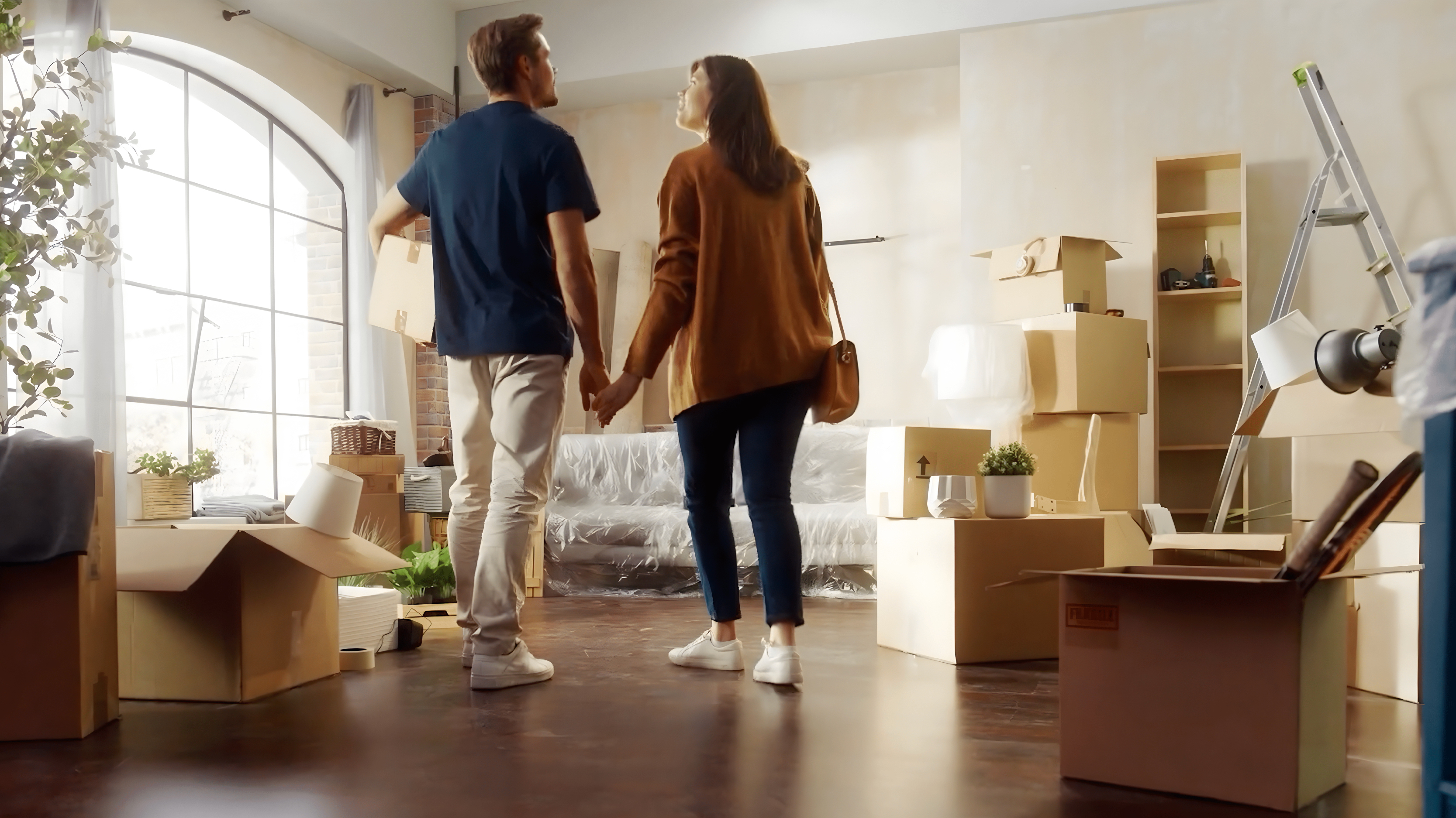 A couple walking hand in hand inside a room filled with moving boxes and furniture, preparing to settle into a new home.