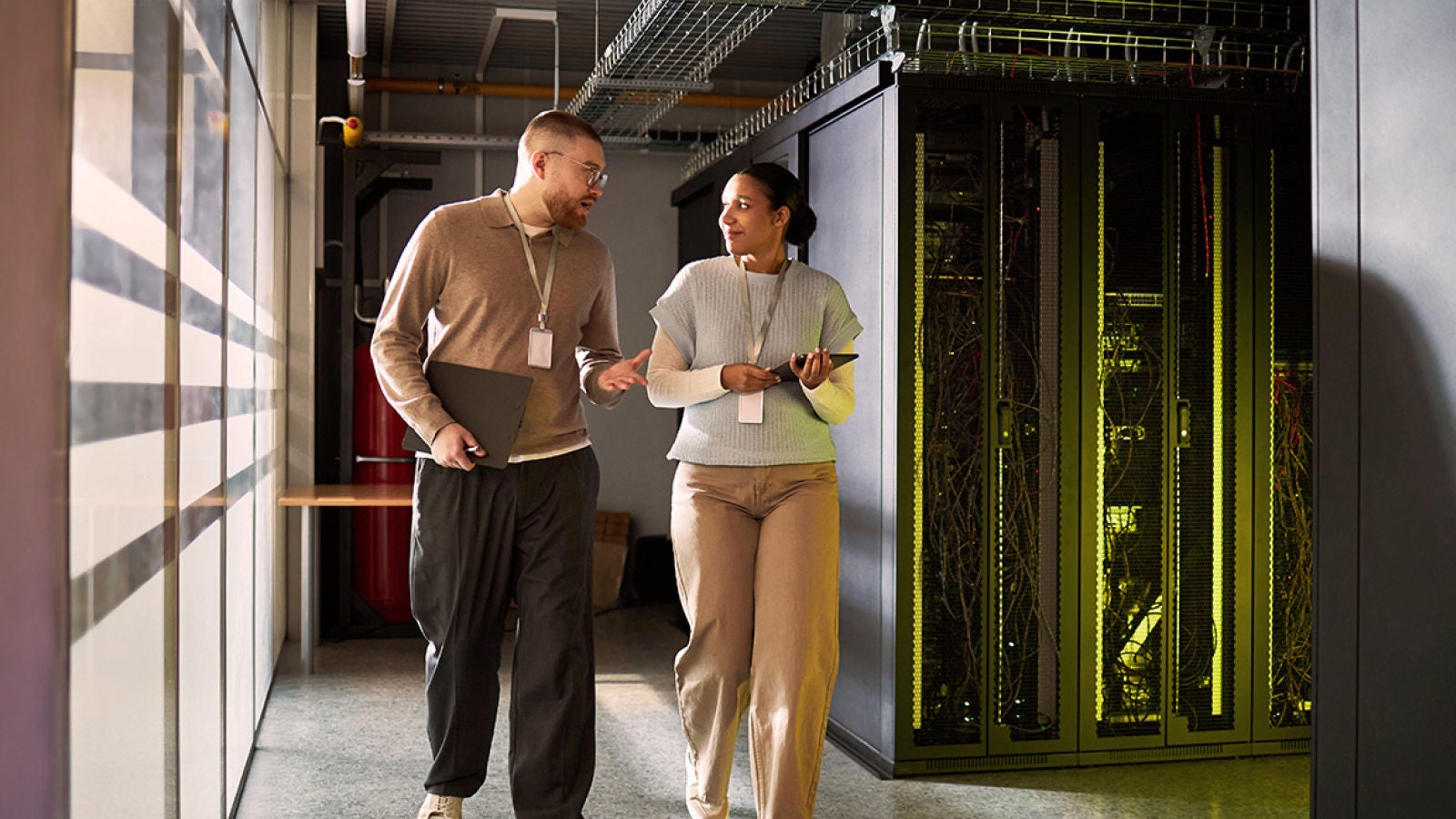 Two technology professionals walk through a data center corridor beside server racks while discussing work.