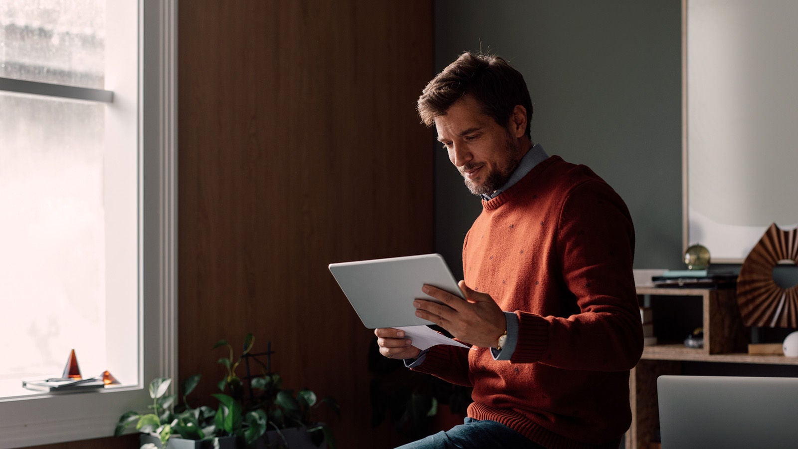 A man in a red sweater sitting by a window in a modern home office, smiling while looking at a tablet.