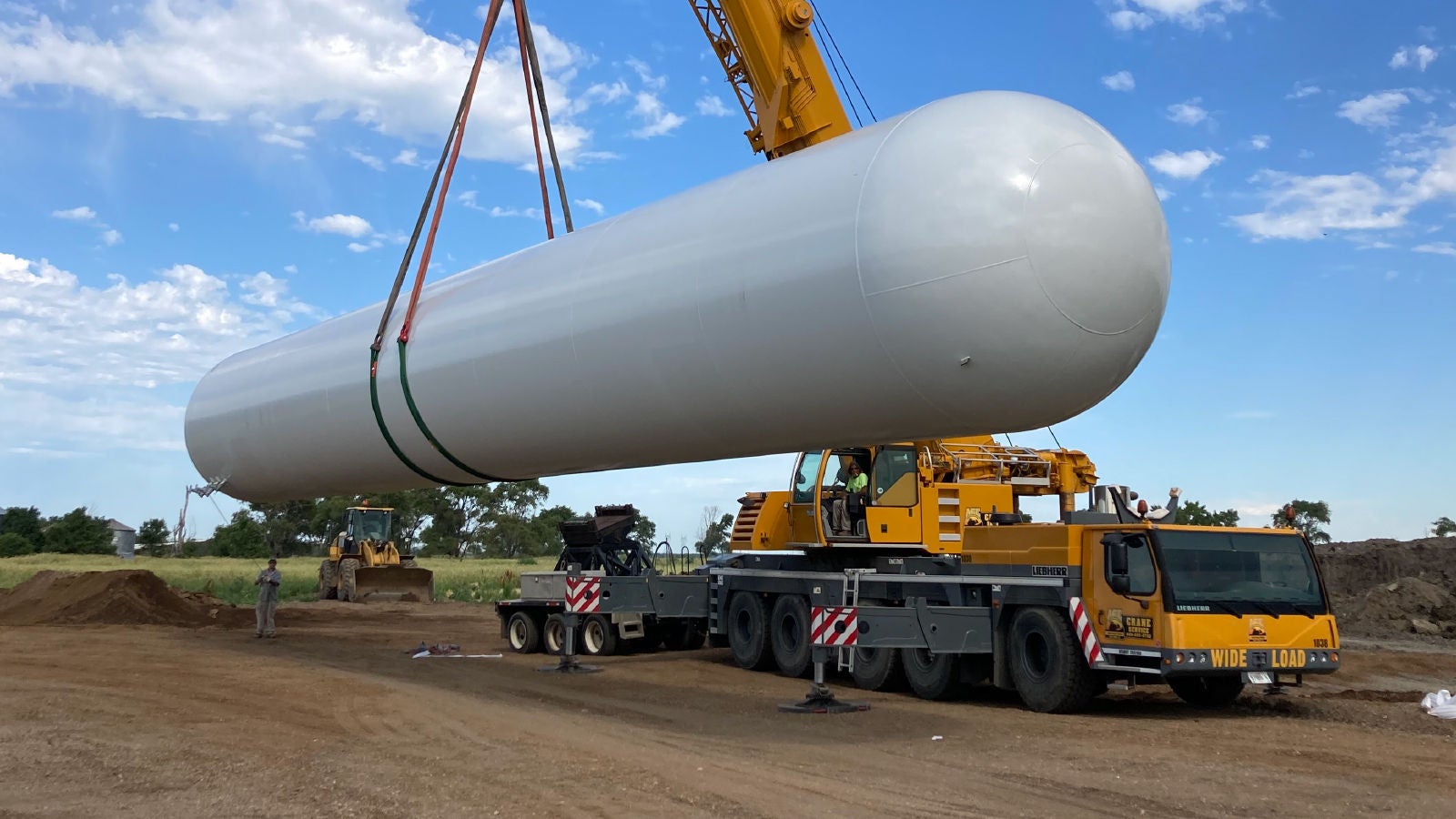 A crane lifting a large cylindrical industrial tank at an outdoor construction site, with workers and heavy machinery visible under a blue sky.