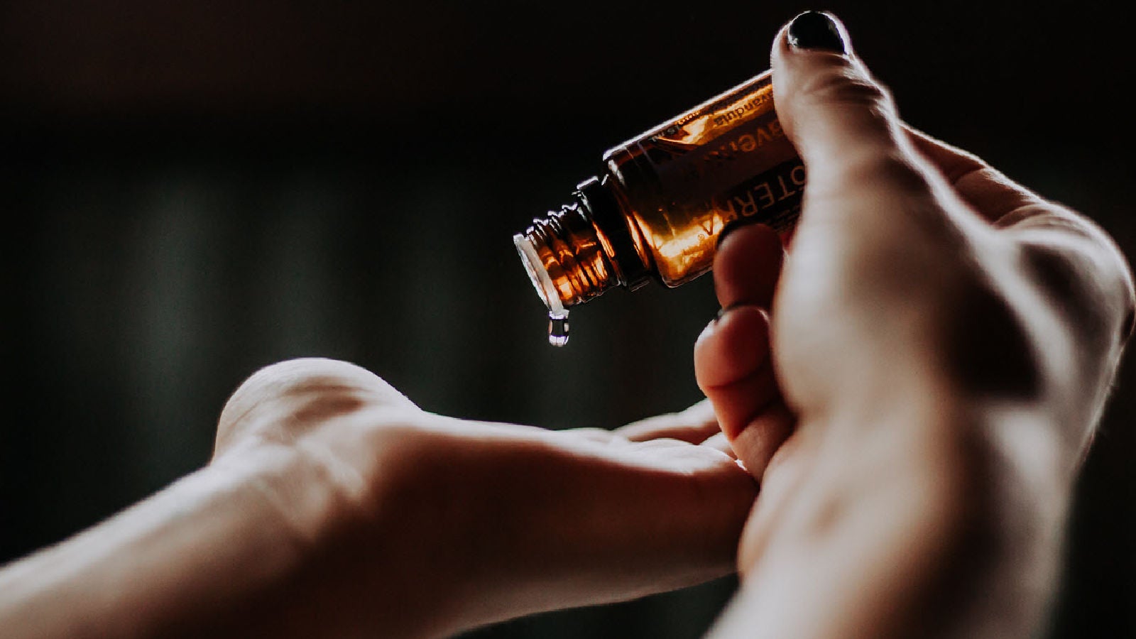 A hand pouring liquid from a small amber essential oil bottle onto a wrist against a dark background.