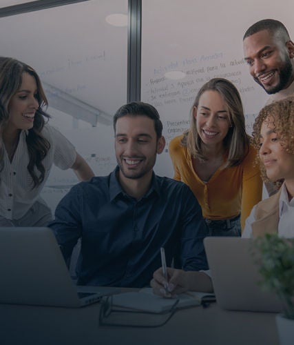 A group of professionals gathered around a laptop in a meeting room, smiling and discussing work with a whiteboard and sticky notes in the background.