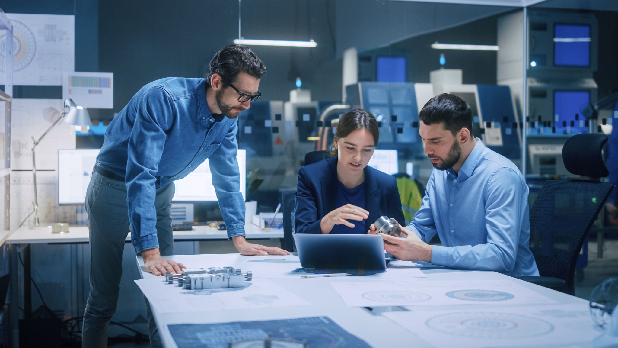 Engineering team in a design studio. Three professionals review technical blueprints and 3D-printed parts around a table, with prototypes in the background.