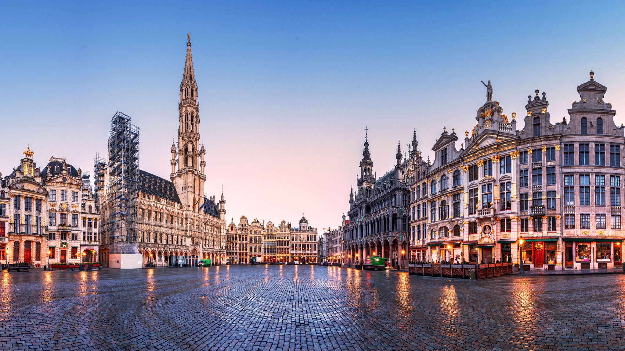 Belgium wide view of a historic European square with ornate guildhalls and a tall central spire, cobblestone ground reflecting warm evening light.