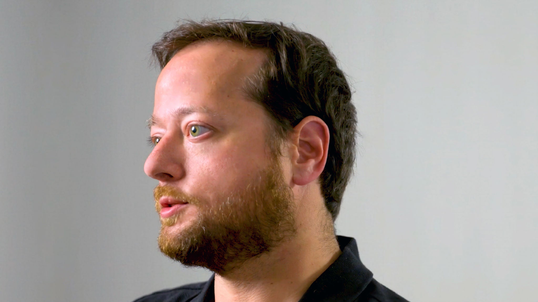 Close-up of a man speaking while looking slightly to the side against a plain background.