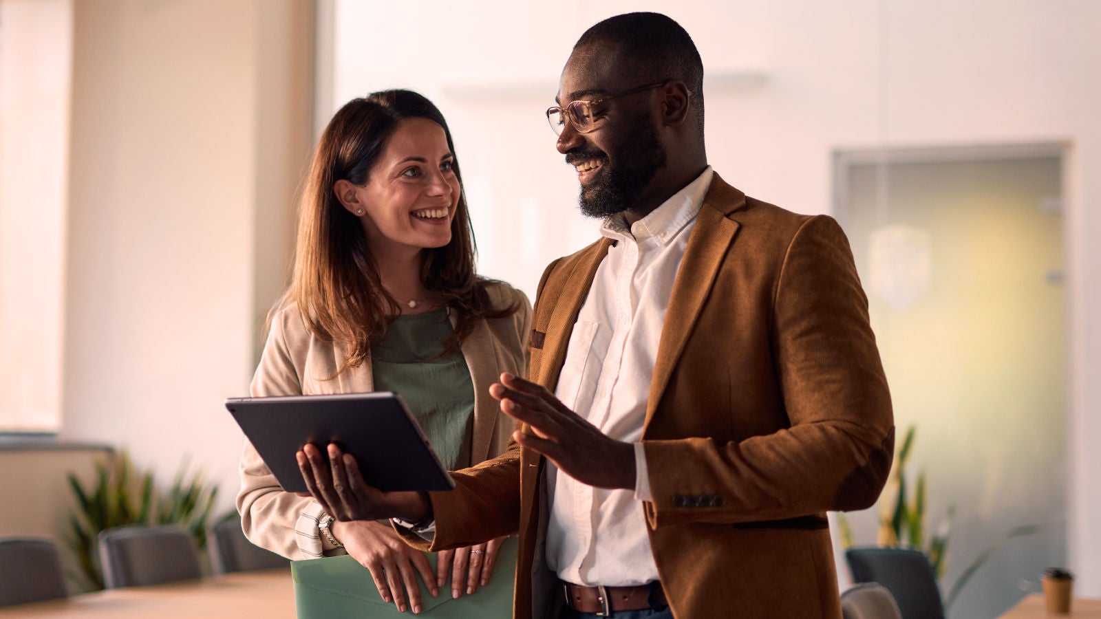 Two colleagues standing in a conference room, smiling and conversing while one holds a tablet.