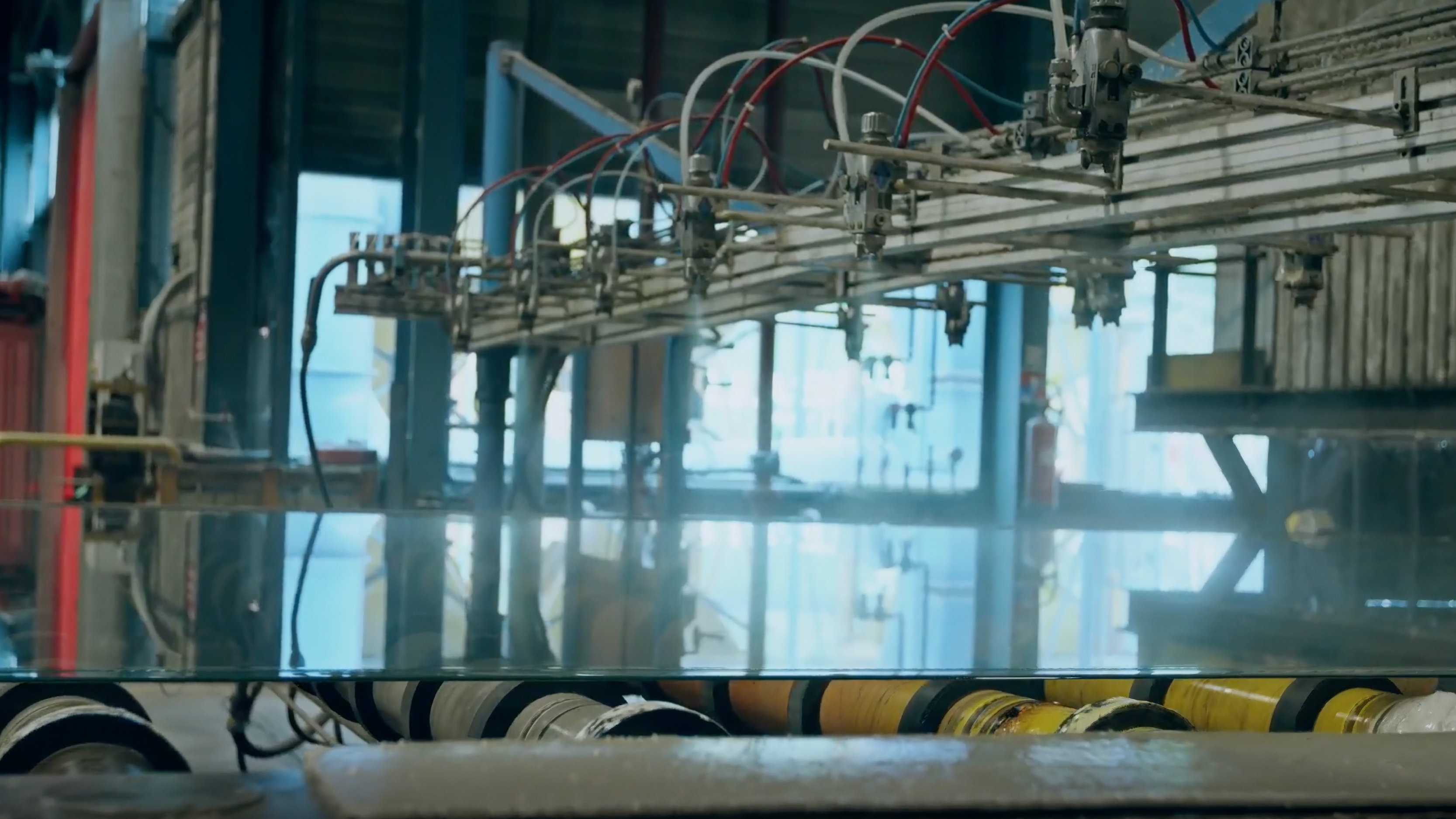 AGC Glass Customer Story Illustrative Image 2 Industrial glass manufacturing equipment with overhead tubing and rollers transporting a flat sheet of glass in a factory setting.