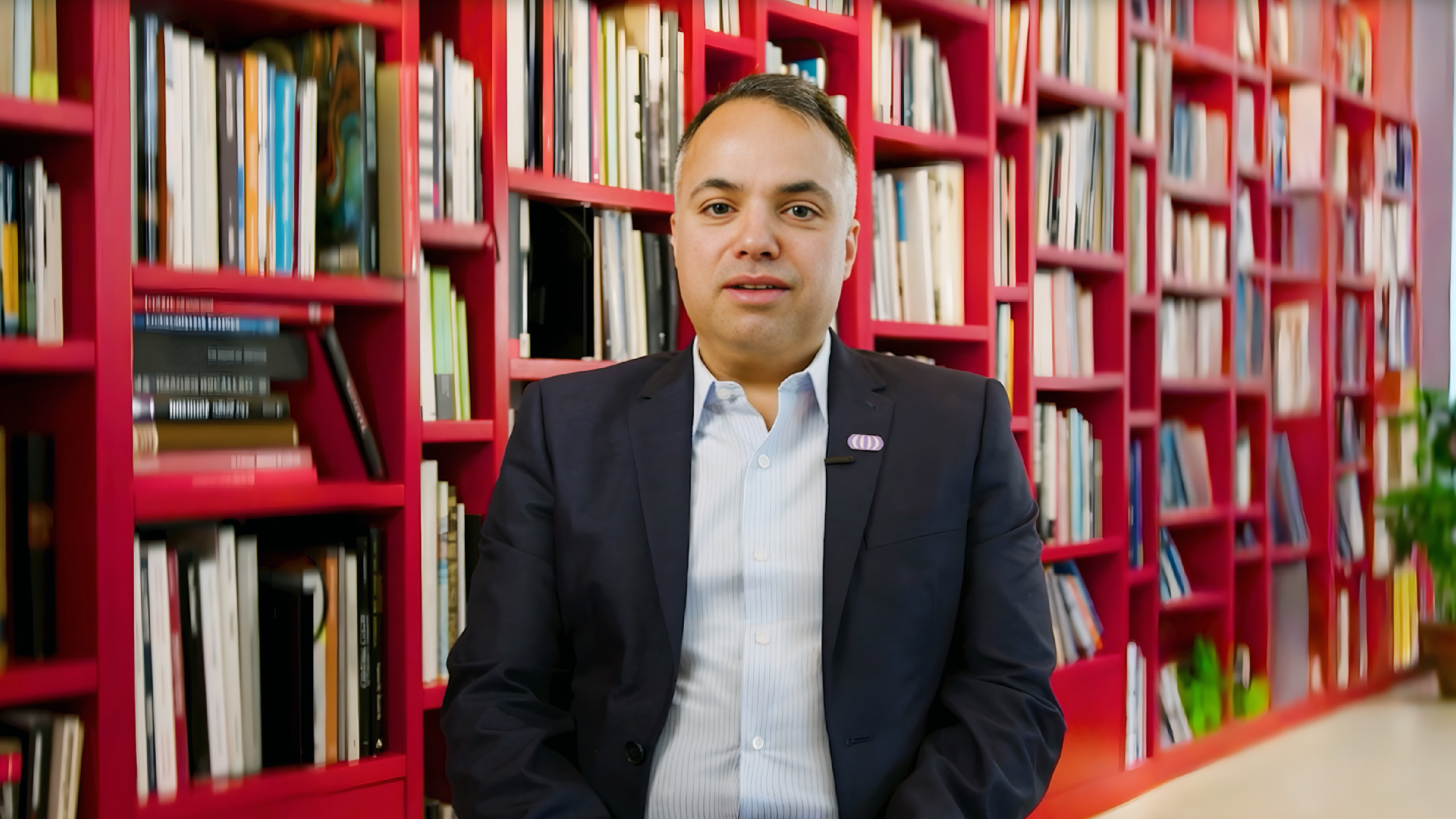 A man seated indoors in front of a red bookshelf filled with books.
