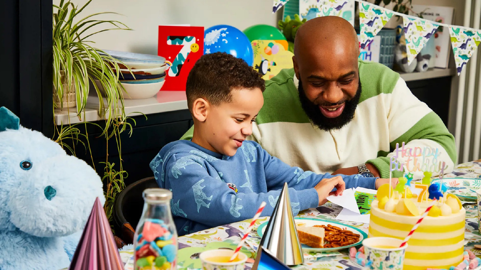 A child celebrating a birthday with an adult at a colorful, dinosaur-themed party table, surrounded by cake, balloons, and decorations.