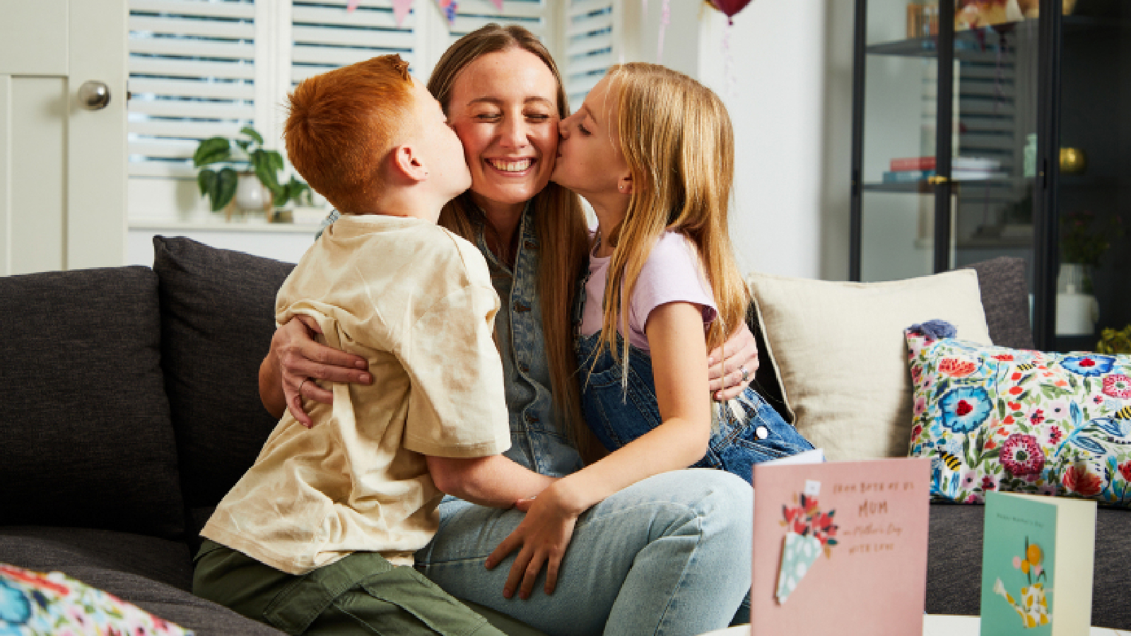 A smiling adult on a sofa being kissed on both cheeks by two children, with greeting cards and colorful pillows visible in the cozy room.