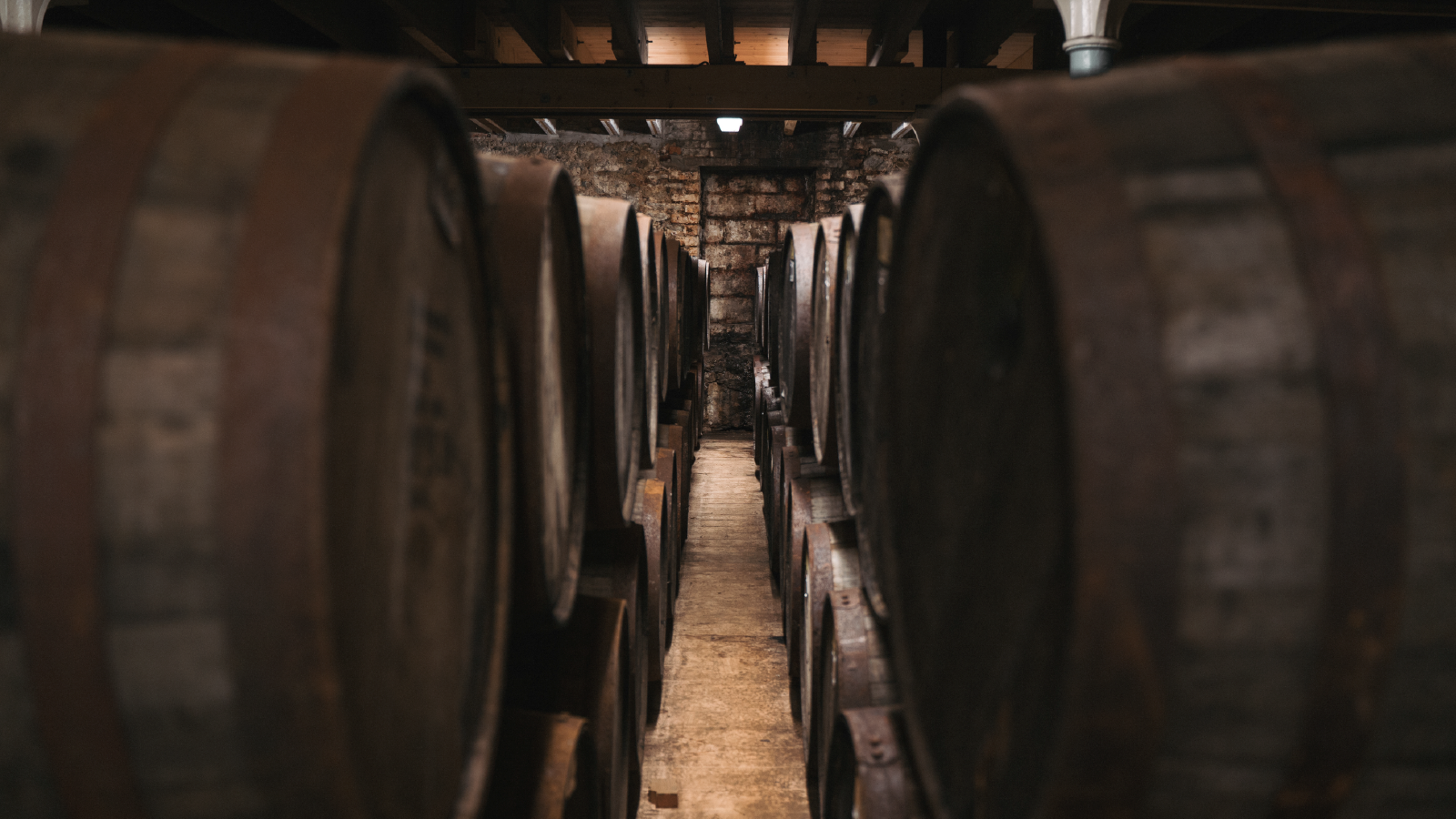 CVH Customer Story Illustrative Image 1 A dimly lit whiskey aging cellar with rows of large wooden barrels stacked on both sides of a narrow aisle.