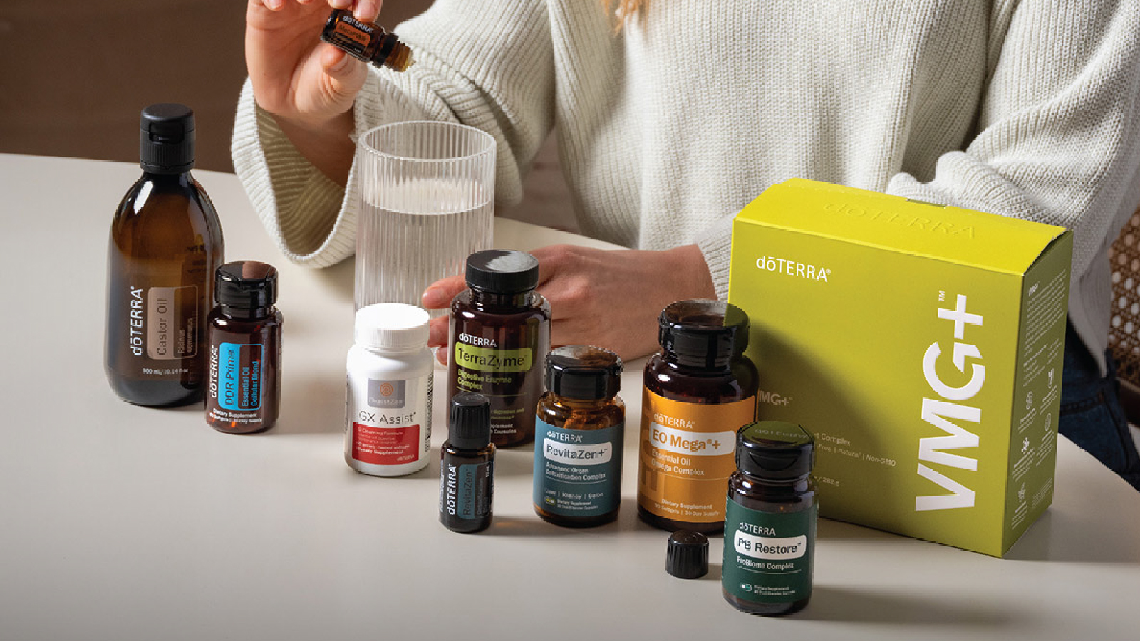 Assorted dōTERRA supplement bottles arranged on a table beside a glass of water and a person holding a small bottle.