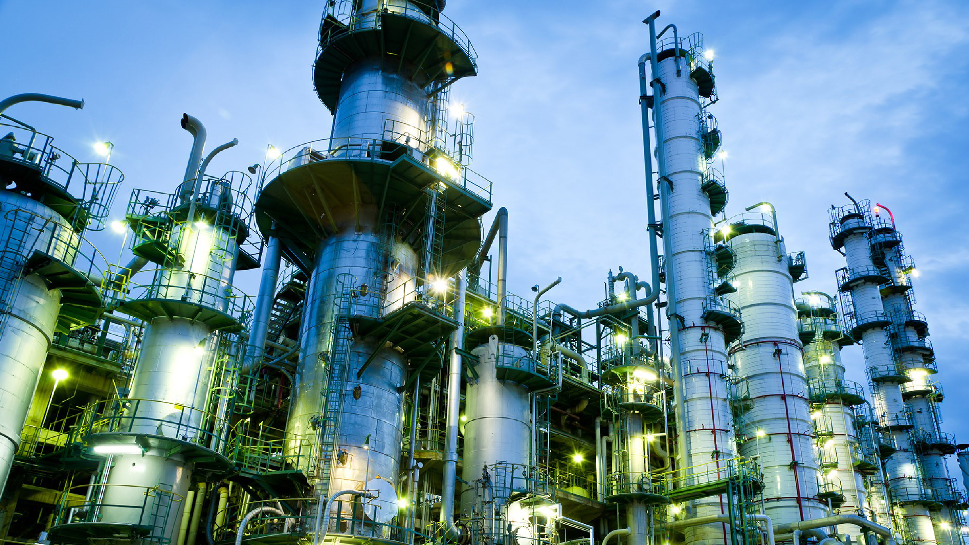 An industrial facility with multiple tall processing towers, pipes, and metal structures illuminated by lights against a blue sky.