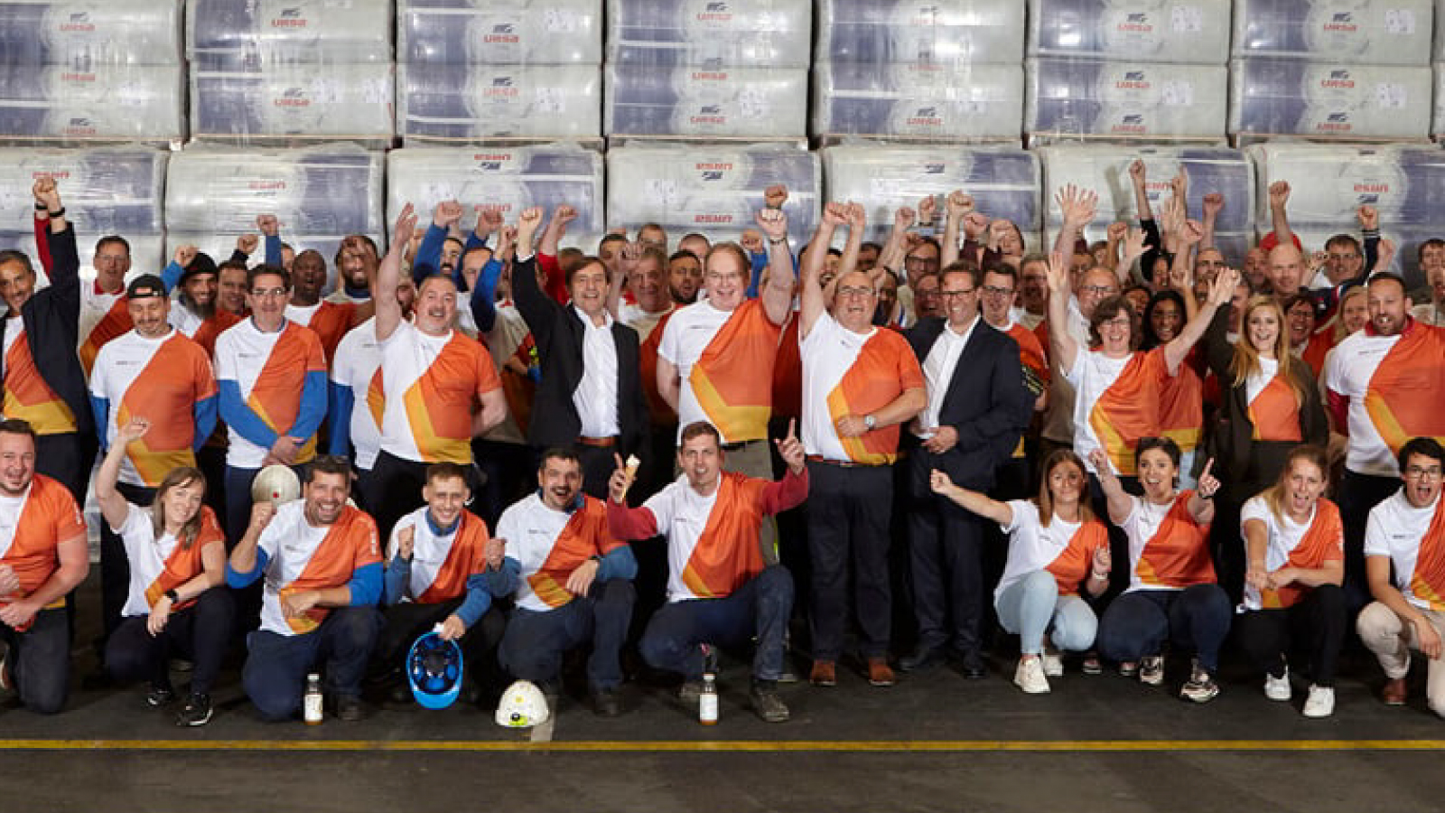 Etex Customer Story Illustrative Image 1 A large group of employees wearing matching orange and white shirts celebrate together in a warehouse, raising their hands in excitement.