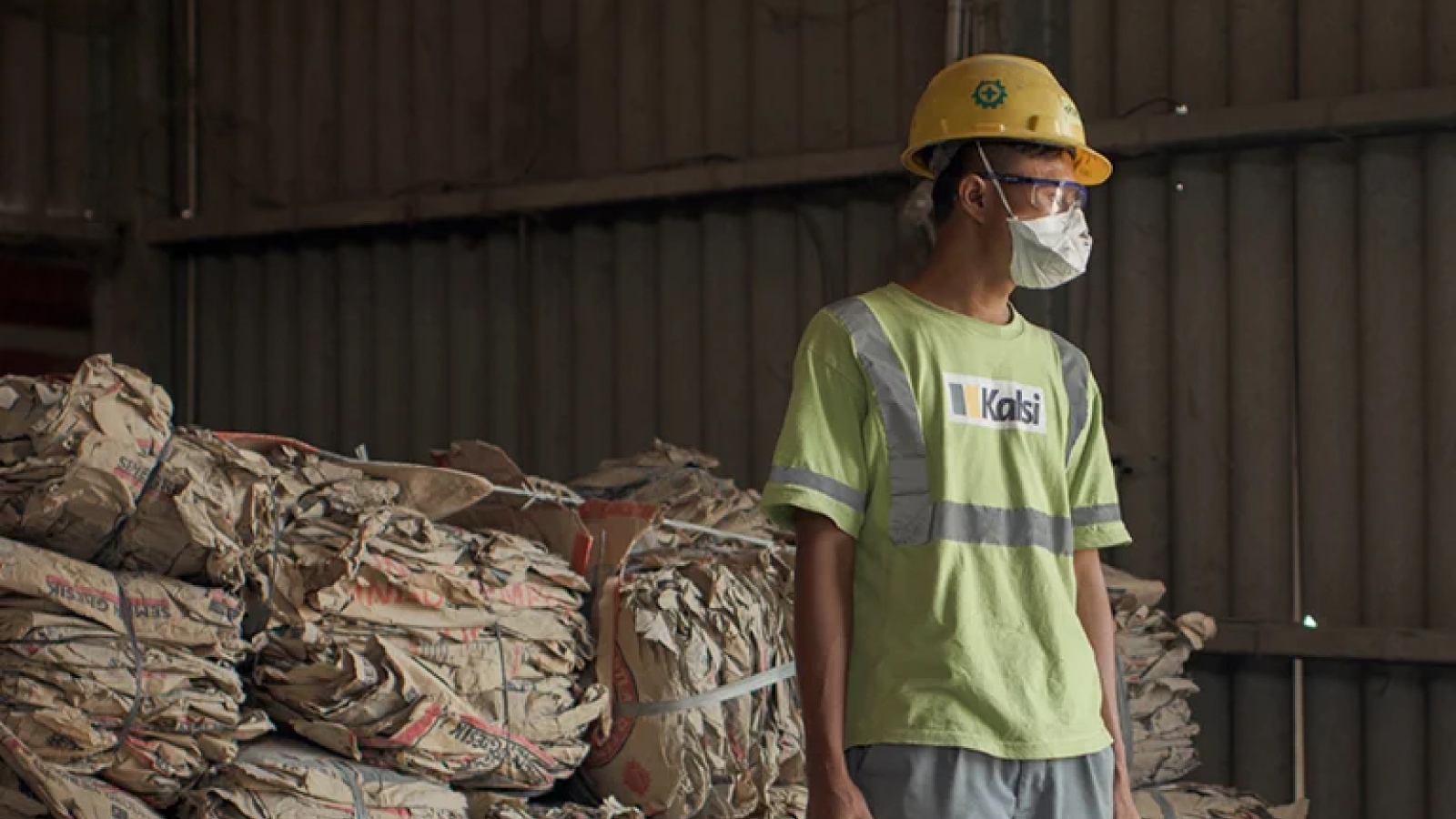 Etex Customer Story Illustrative Image 2 A worker in a green shirt and yellow hard hat stands in a warehouse with stacks of compressed paper packaging behind him. He wears a face mask and safety goggles.