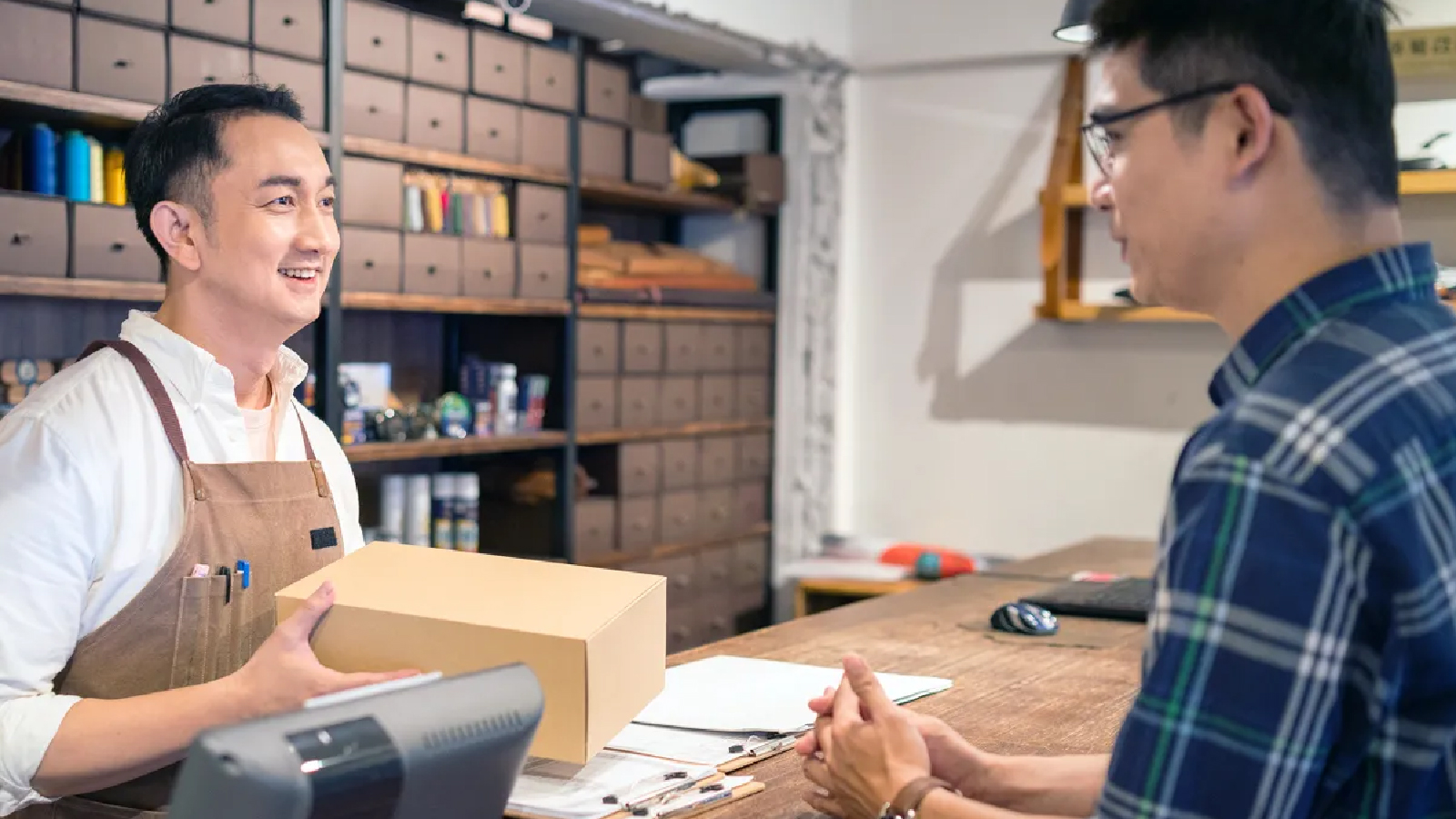 Ingenico Customer Story Illustrative Image 2 A smiling store worker wearing an apron hands a package to a customer across a wooden counter in a shop with neatly organized drawers.