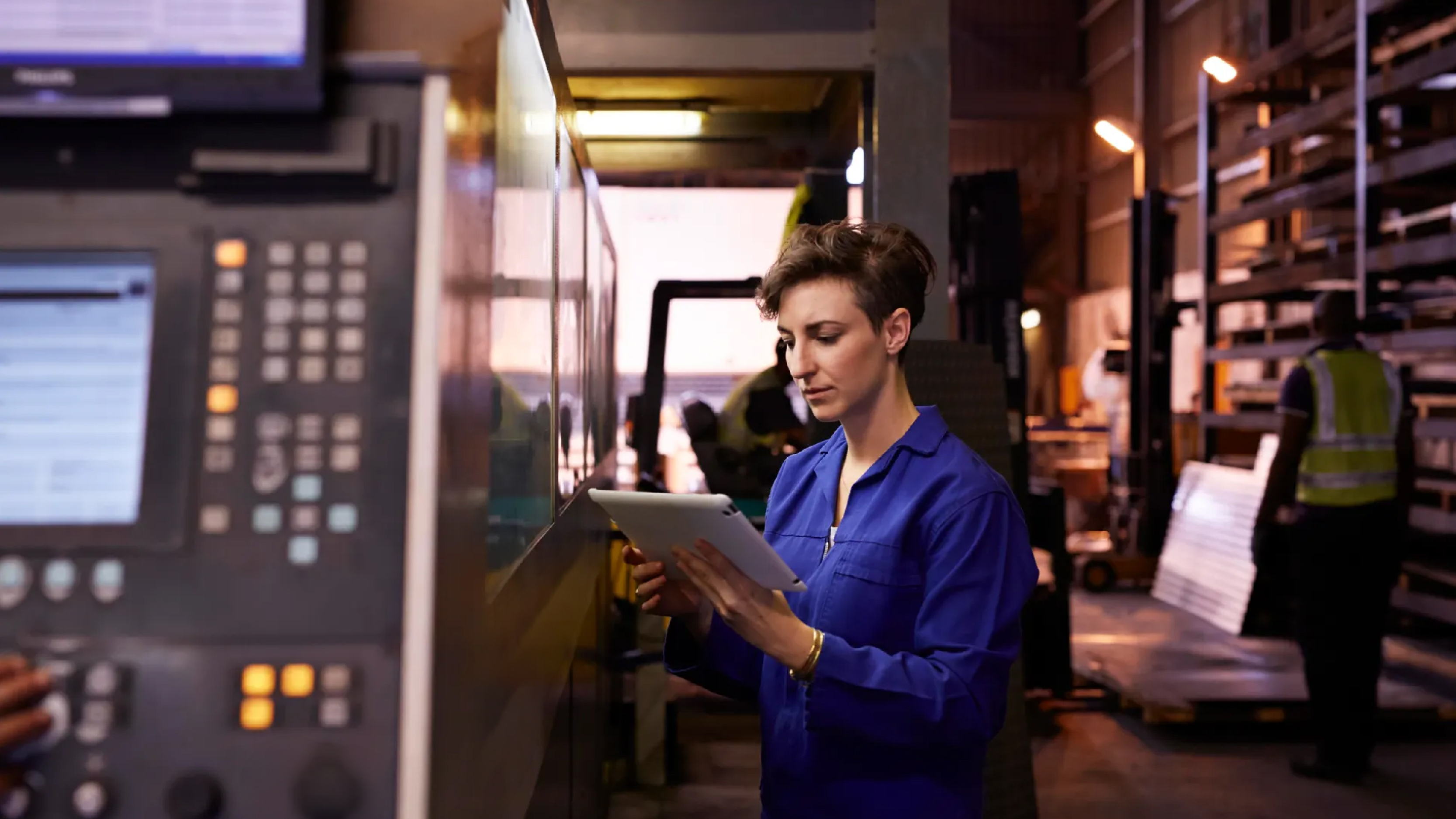 A factory worker in blue coveralls uses a tablet beside industrial machinery on a manufacturing floor, with equipment panels and warehouse activity in the background.