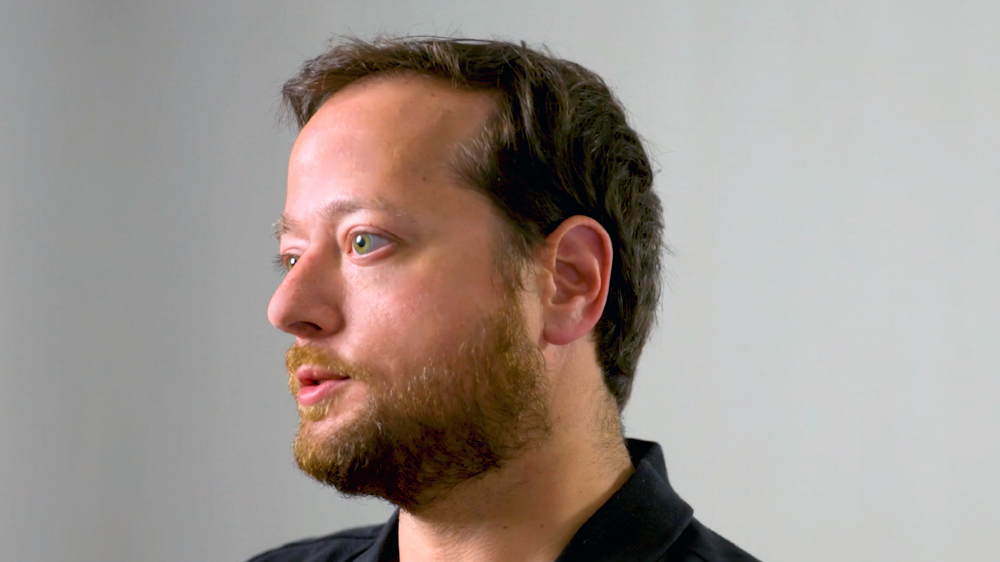 Close-up of a man speaking while looking slightly to the side against a plain background.