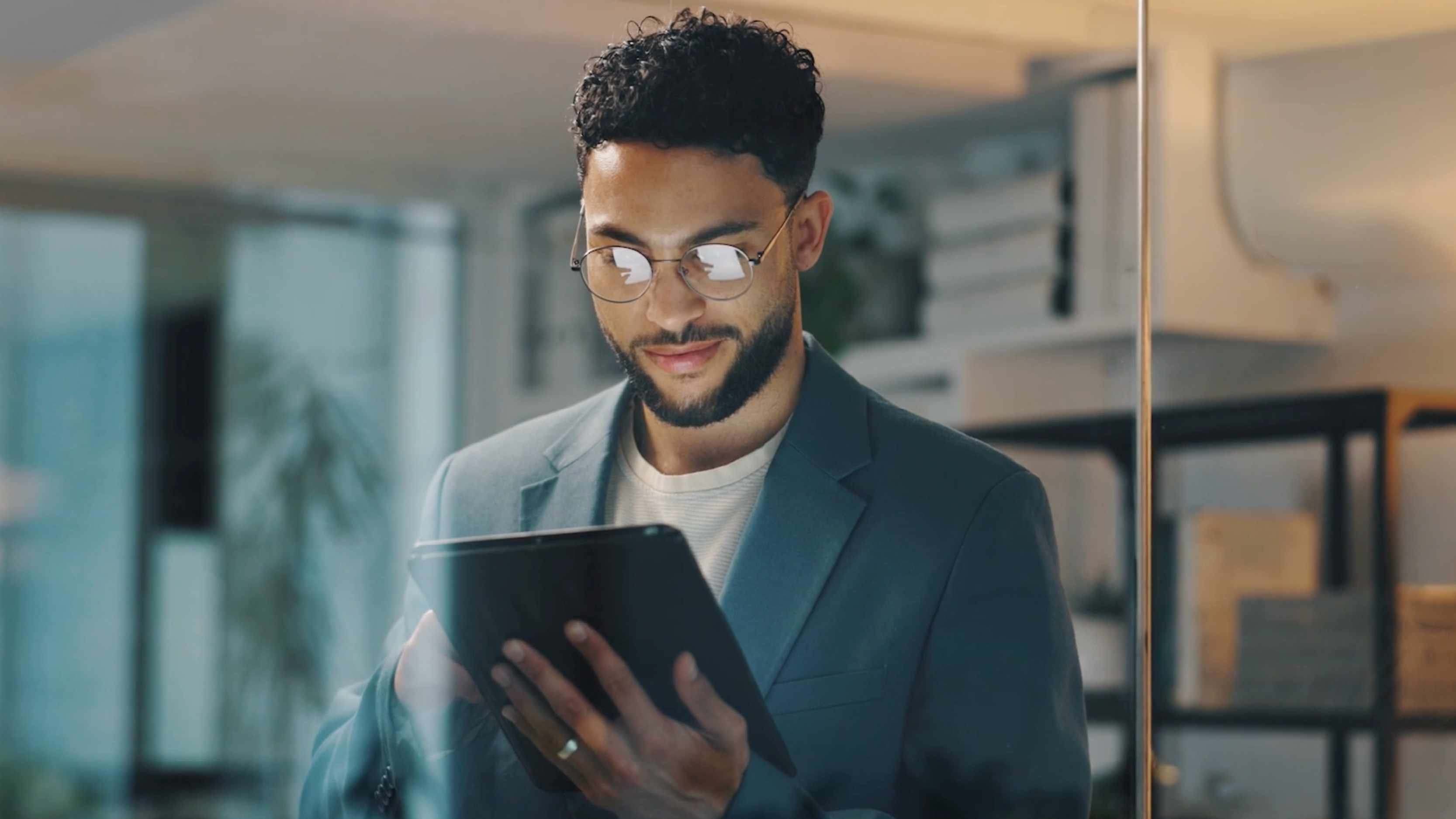 A man wearing glasses reviewing content on a tablet while standing in a modern office space.