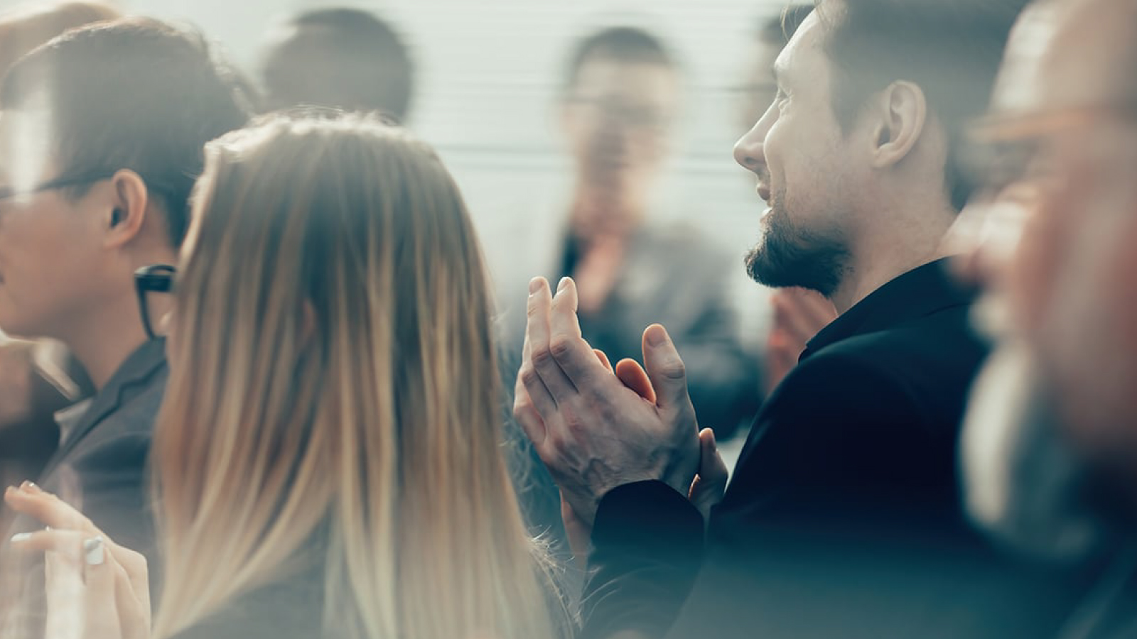 QIC Customer Story Illustrative Image 1 Group of professionals clapping in a softly lit room, with blurred foreground elements and a focus on engaged expressions and applause.
