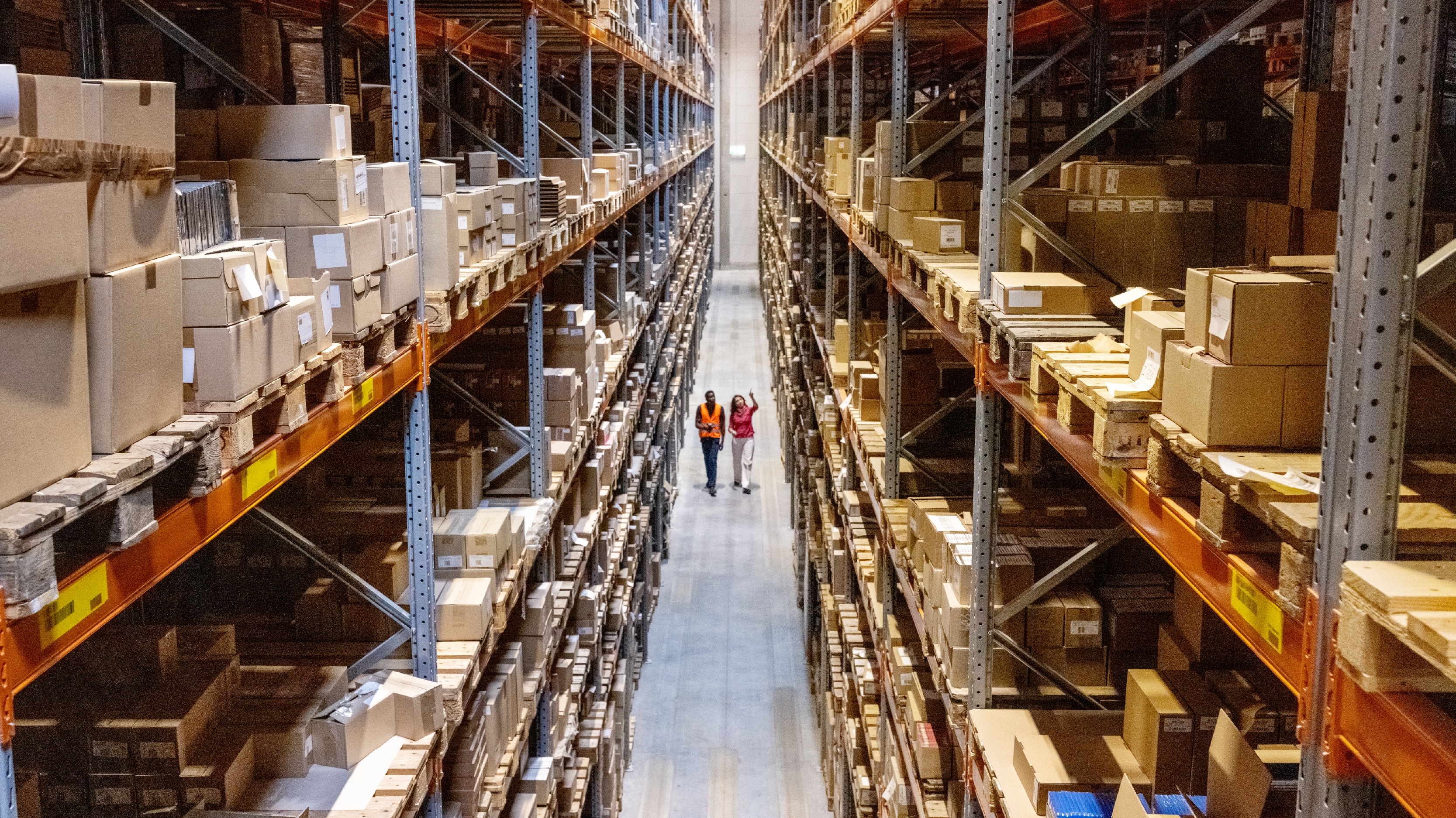 A wide view of a warehouse aisle stacked with rows of boxes as two people walk together down the center walkway.