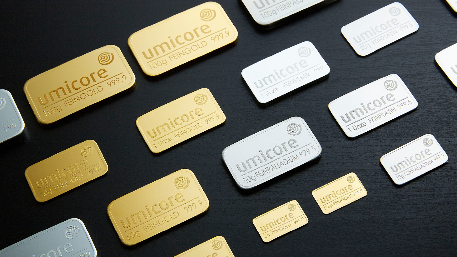 Umicore Customer Story Illustrative Image 1 A variety of gold, silver, platinum, and palladium bars branded with the Umicore logo are arranged on a black surface, showcasing different weights and metal types.