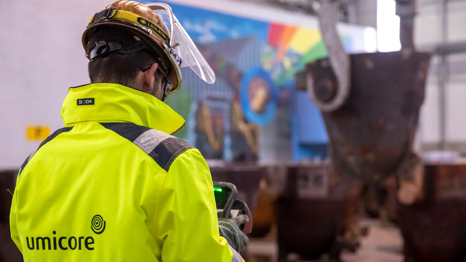 Umicore Customer Story Illustrative Image 2 A worker in a high-visibility yellow jacket and hard hat operates a handheld device inside an industrial facility, with large metal containers and a colorful mural visible in the background.