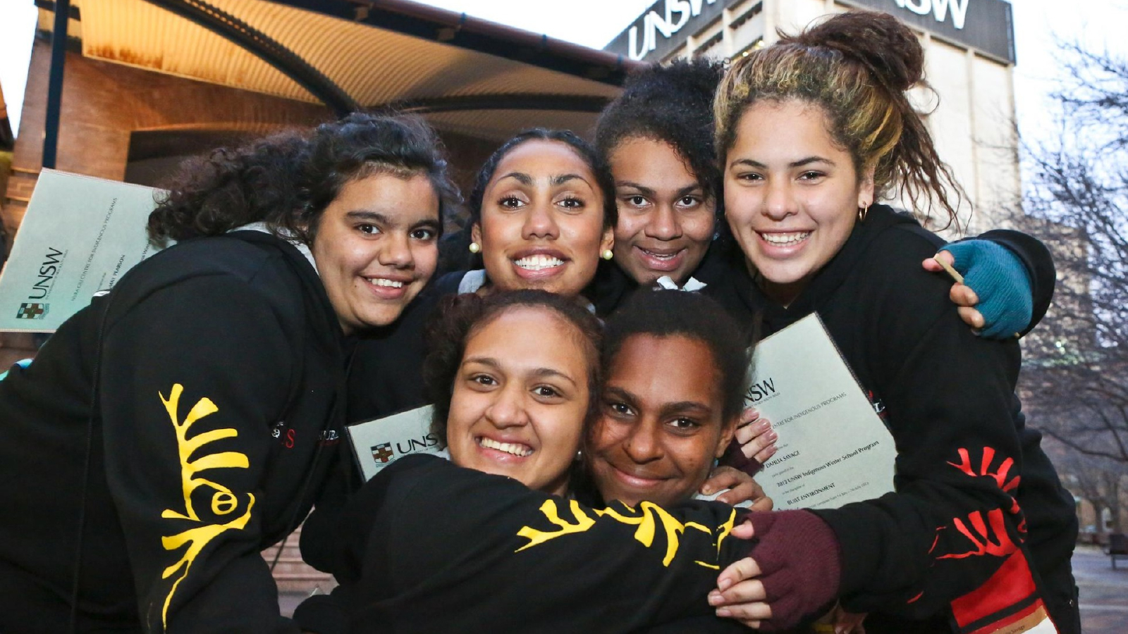 A group of six smiling students hugging closely outdoors, holding UNSW Sydney folders.