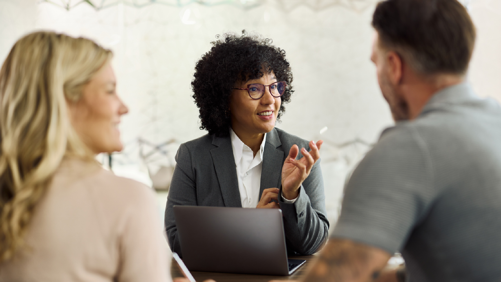 A financial advisor with curly hair and glasses sits at a table with an open laptop, speaking and gesturing with her hand while consulting a couple seated across from her.