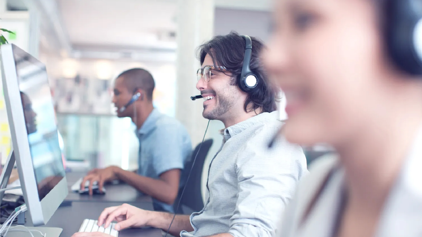 Ingenico Customer Story Illustrative Image 1 A smiling man wearing a headset works at a computer, with other colleagues in the background.
