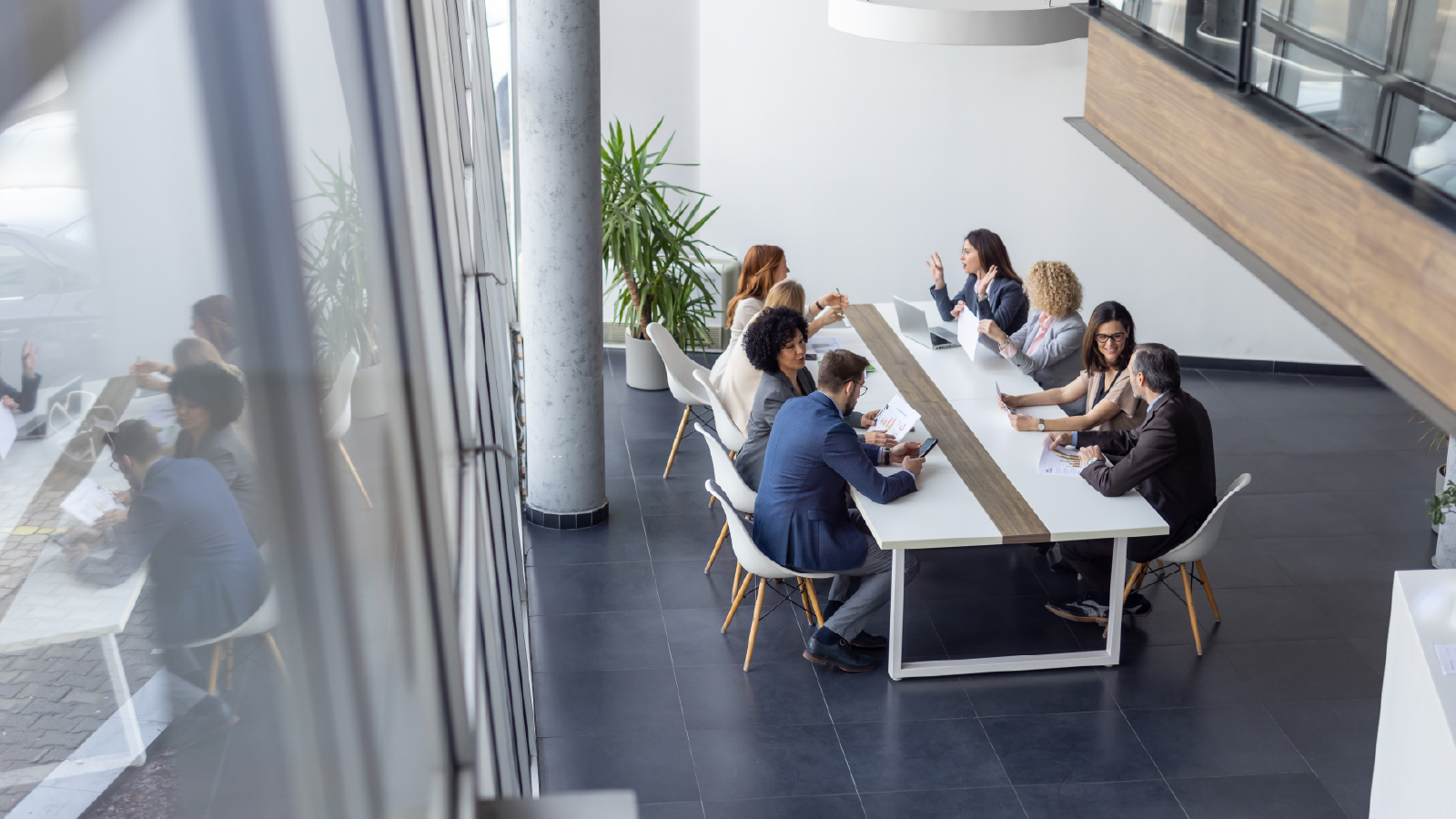 A large group of colleagues sit around a long white conference table in a bright, modern office space, engaged in discussion and reviewing documents. Tall windows reflect the scene, adding depth to the image.