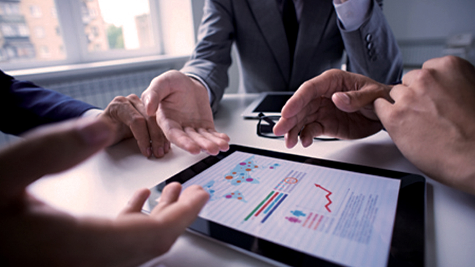 Nitto Denko Customer Story Illustrative Image 1 A close-up of a business meeting where hands are gesturing over a tablet displaying graphs and charts. The scene suggests a financial or strategic discussion.
