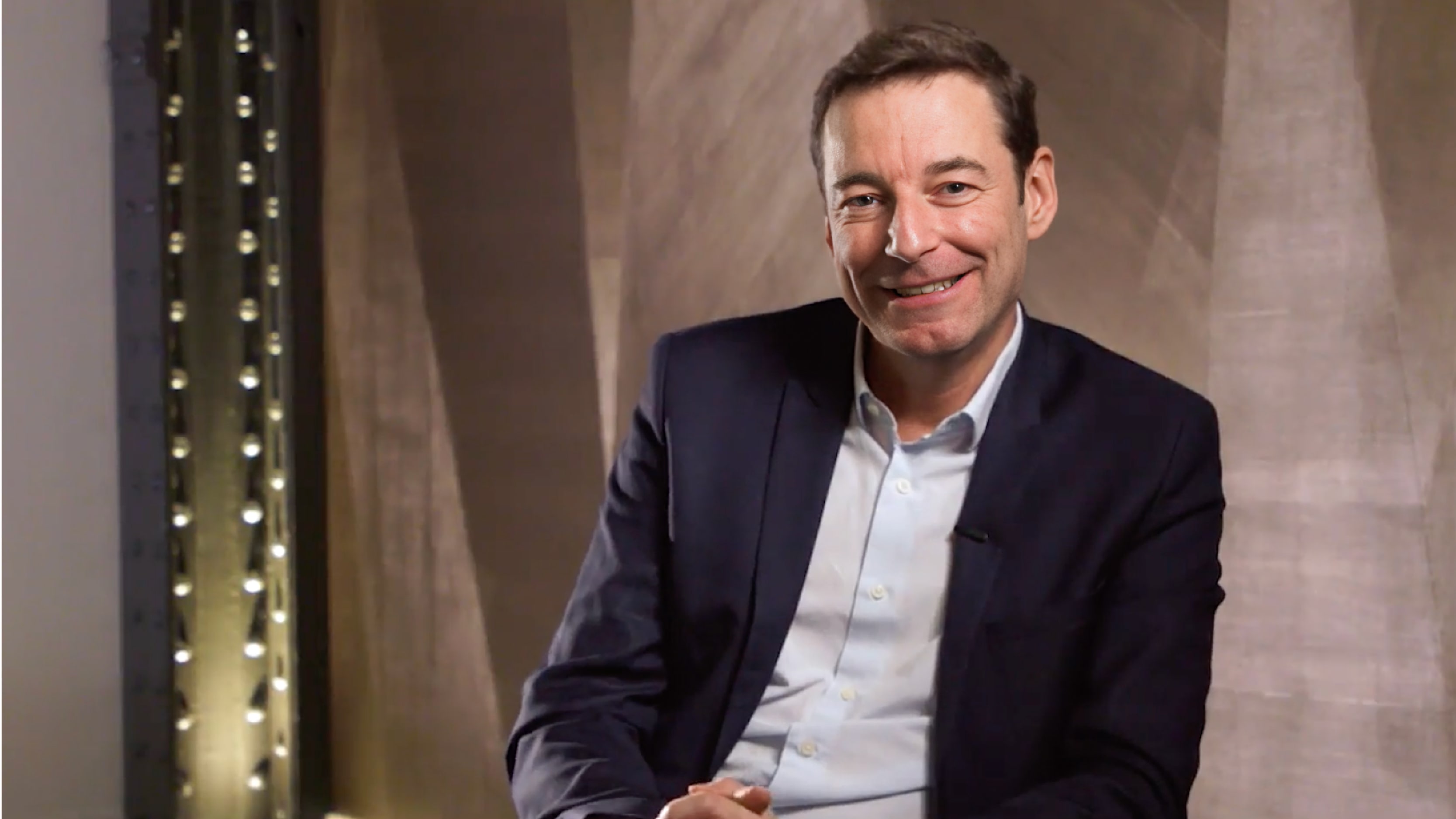 A man in a suit smiling while seated against a softly lit, modern brown backdrop with vertical lighting accents.