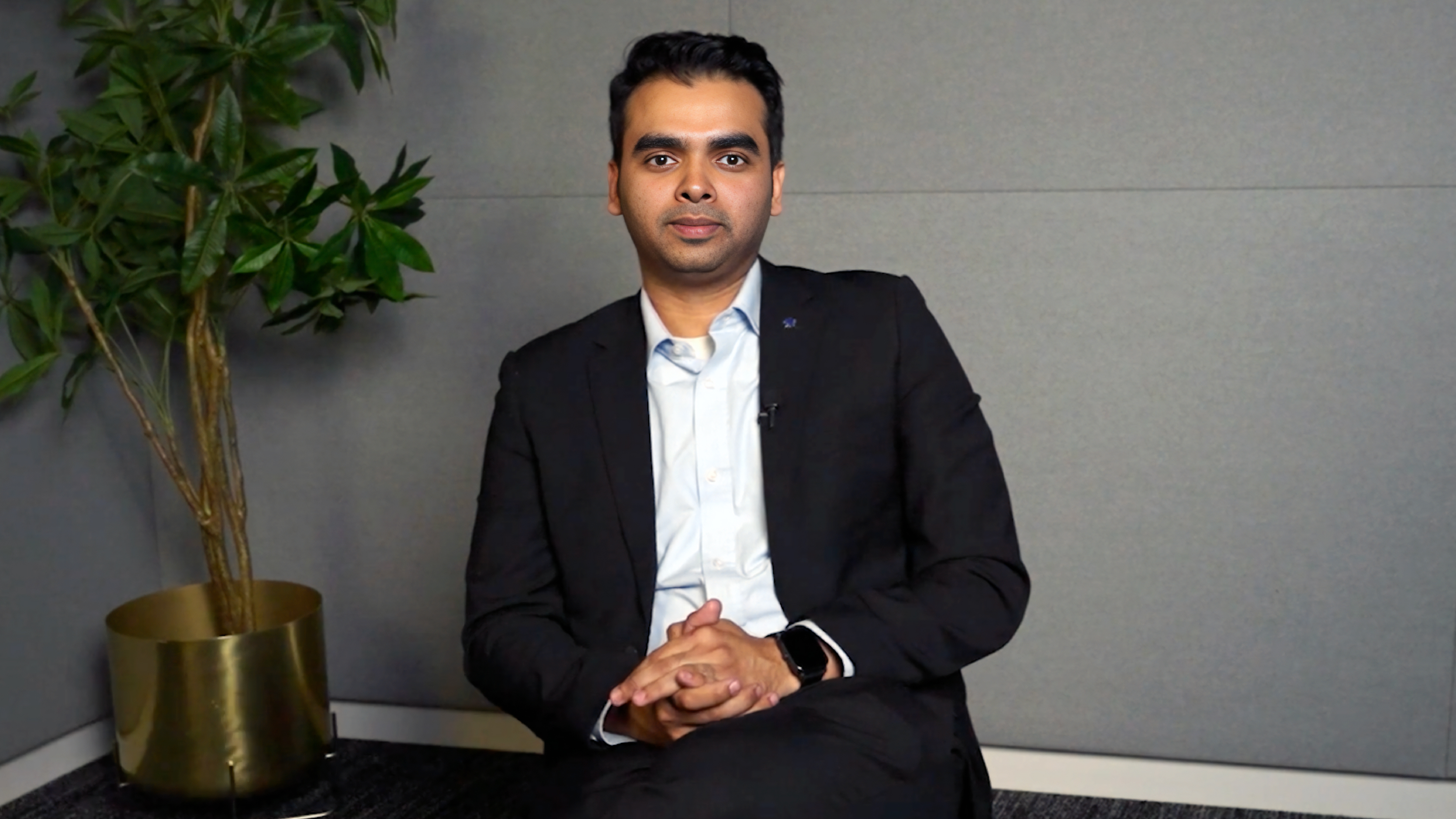 A man in a suit seated in an office setting with a potted plant beside him, looking directly at the camera.