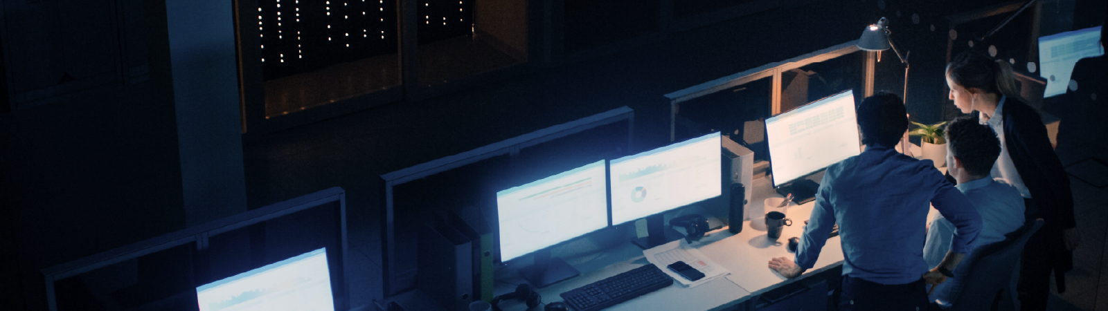Three professionals in a dimly lit office working together at a desk with multiple glowing monitors.