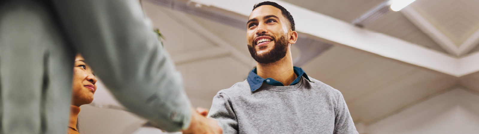A smiling man in a gray sweater shakes hands with another person in a professional setting, with a woman in an orange sweater observing in the background. The ceiling has exposed beams and bright lighting.