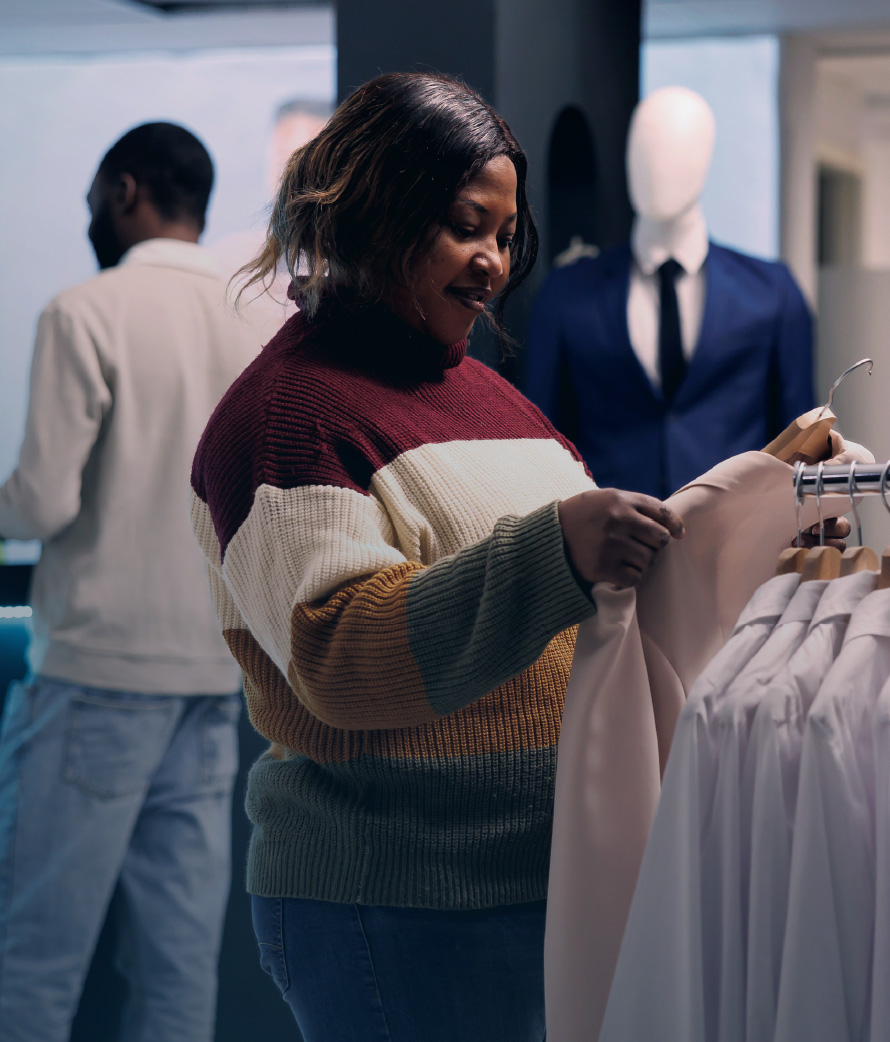 A woman reviewing garments on a clothing rack in a modern retail environment, with folded shirts, mannequins, and other shoppers visible in the store interior.