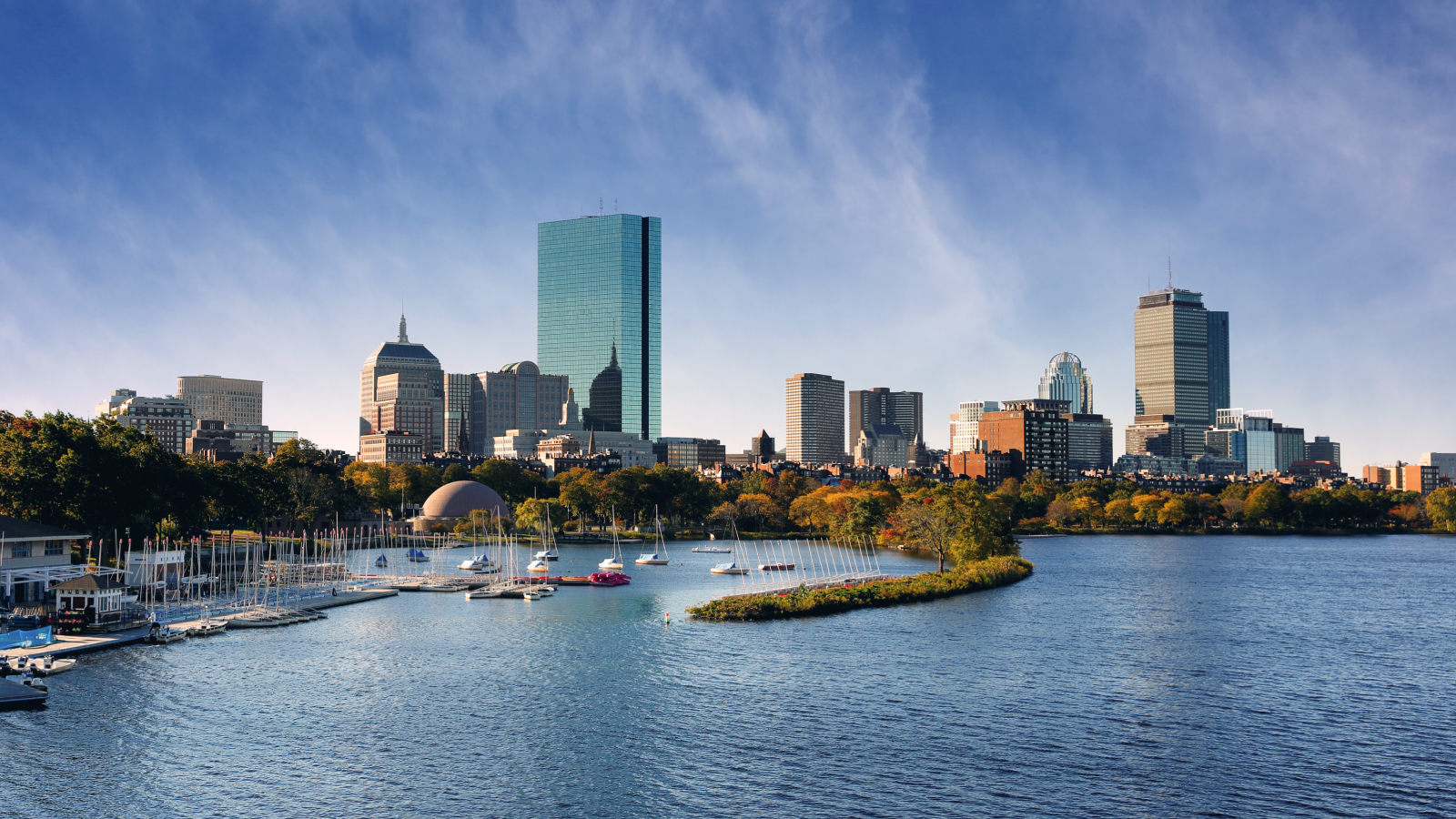 View of the Boston skyline with sailboats docked on the Charles River under a clear blue sky, featuring notable buildings such as the Prudential Tower and John Hancock Tower.