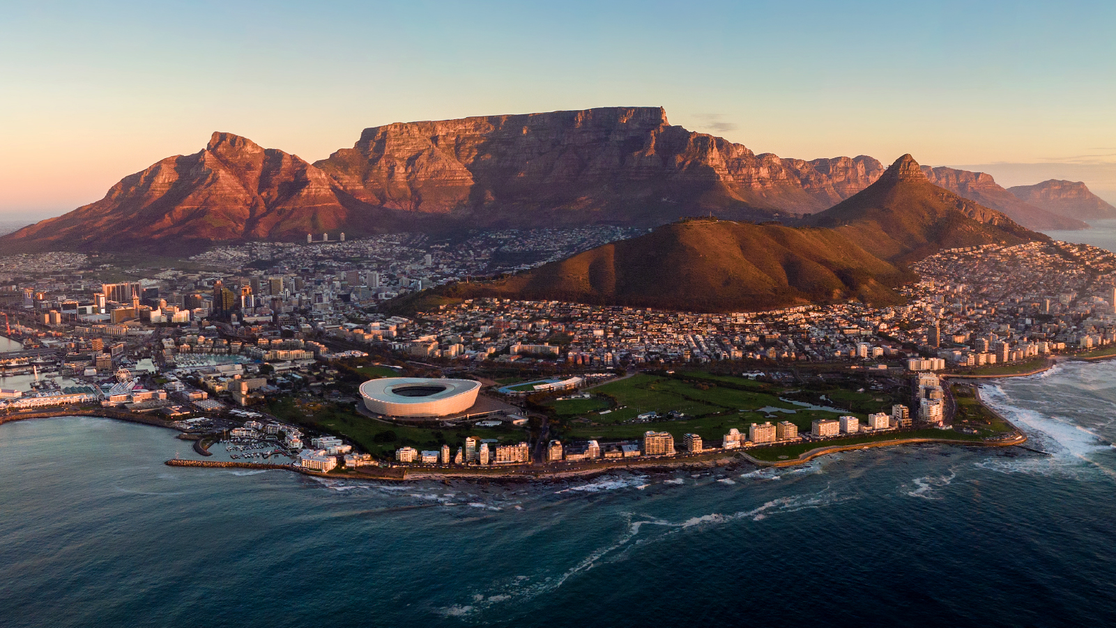Aerial view of Cape Town, South Africa, showing the coastline, Cape Town Stadium, and Table Mountain at sunset.