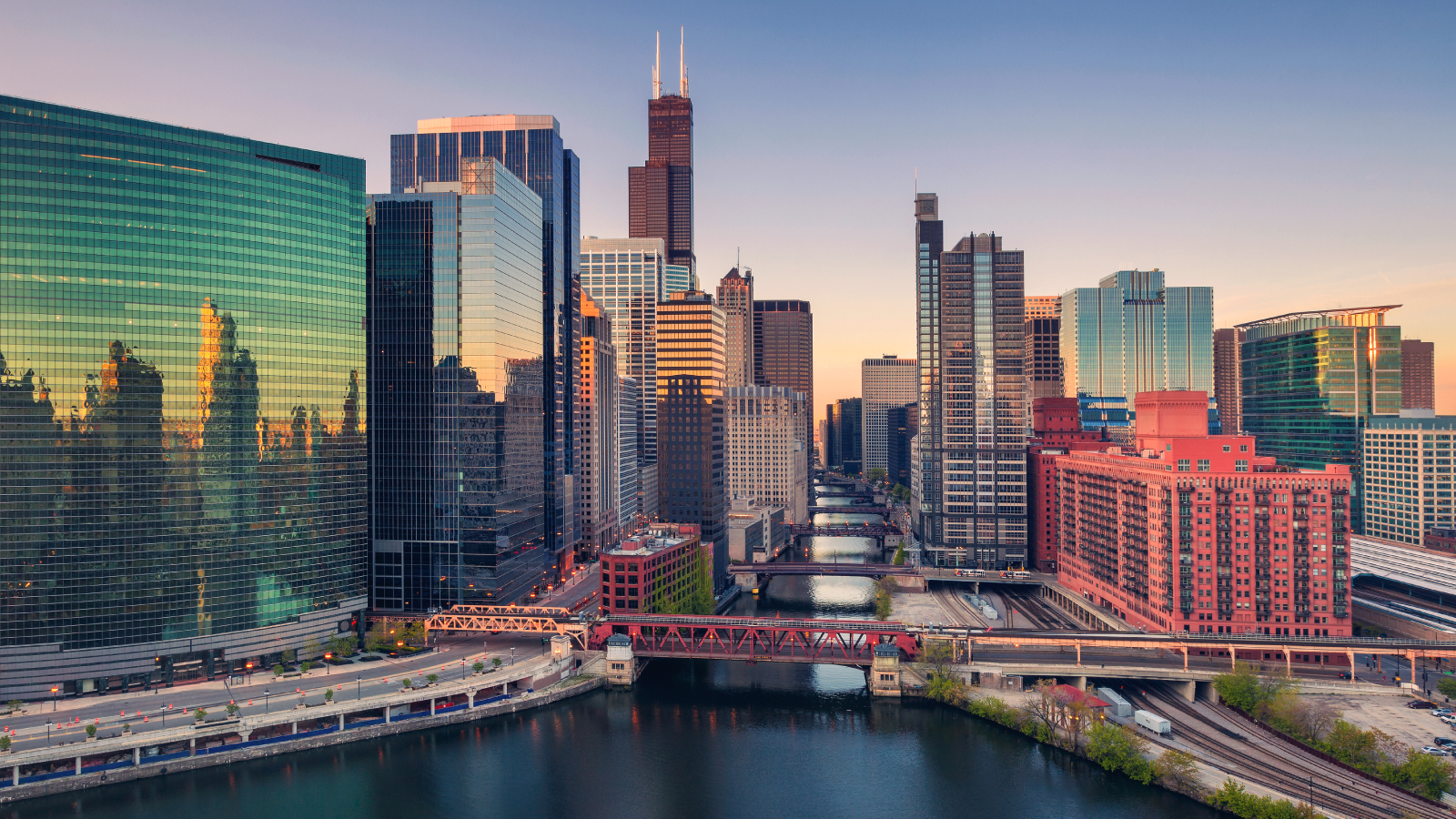 View of the Chicago skyline at sunset featuring the Willis Tower, glass skyscrapers, and red bridges spanning the Chicago River under a clear sky.