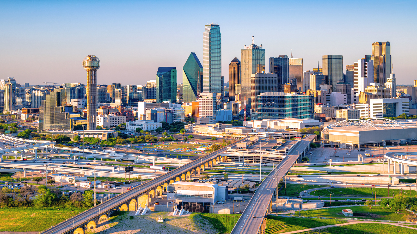 View of the Dallas skyline at sunset featuring the Reunion Tower, modern skyscrapers, and a network of highways in the foreground under a clear sky.