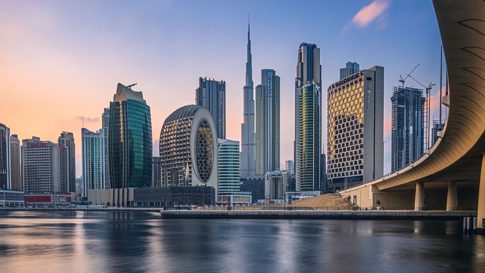 Panoramic view of Dubai skyline featuring Burj Khalifa and surrounding skyscrapers at sunset. The oval-shaped hotel tower and modern high-rises stand along the waterfront, with reflections on the calm water and a bridge on the right side.