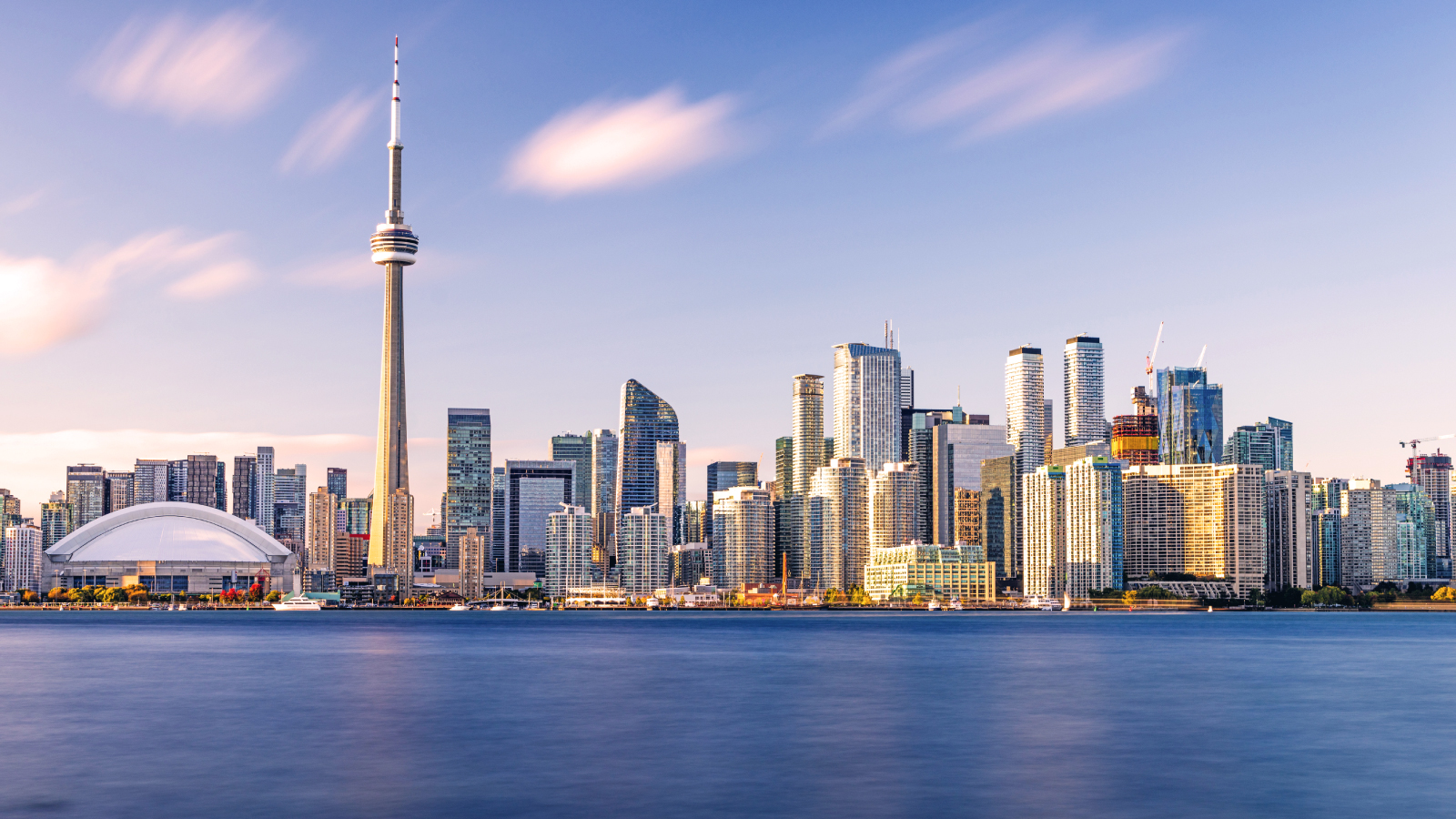 View of the Toronto skyline at sunset featuring the CN Tower, Rogers Centre, and surrounding high-rise buildings reflected in the calm waters of Lake Ontario.