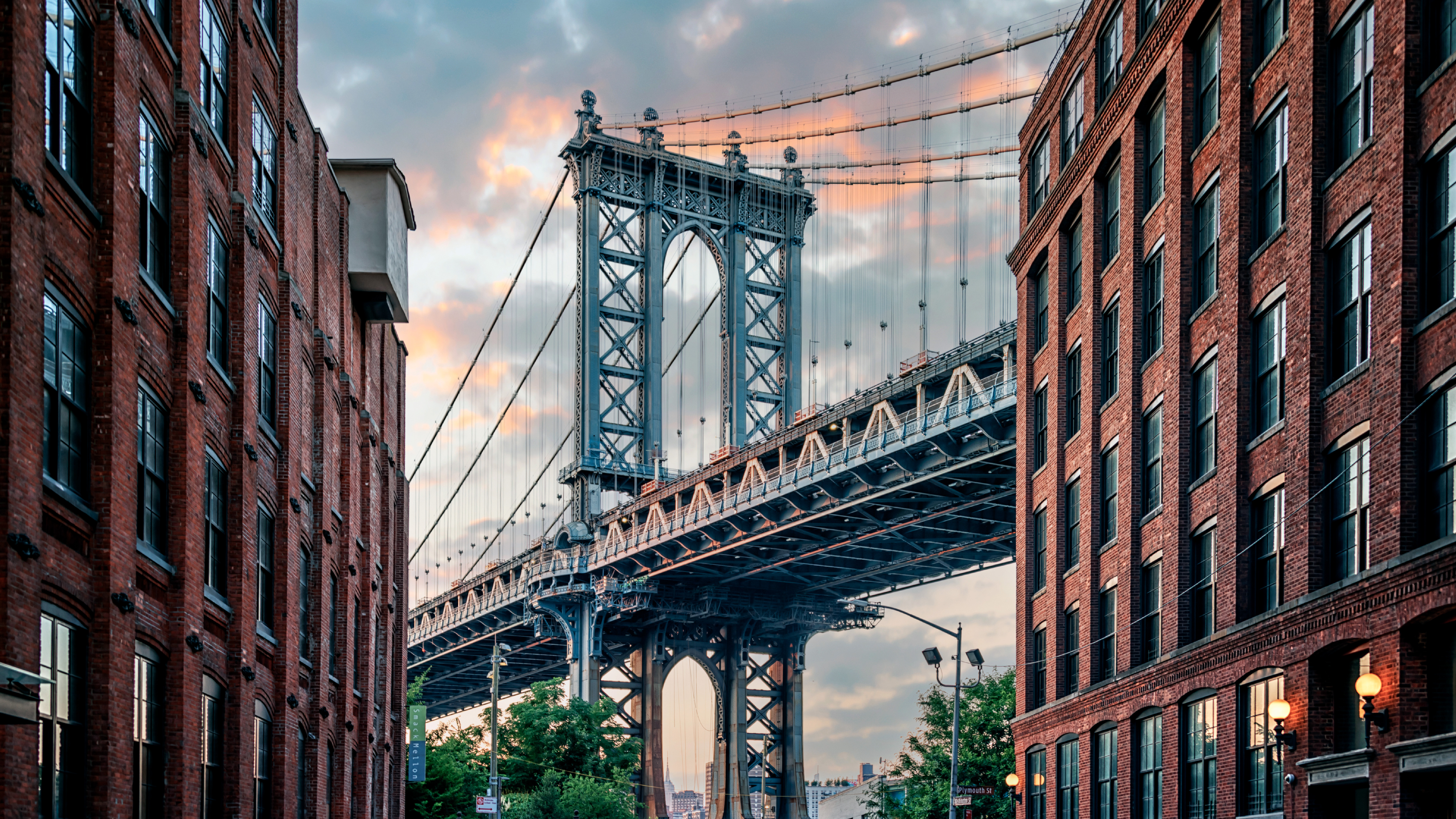 The Manhattan Bridge framed between red brick buildings at street level during sunset, with steel cables and city details visible.