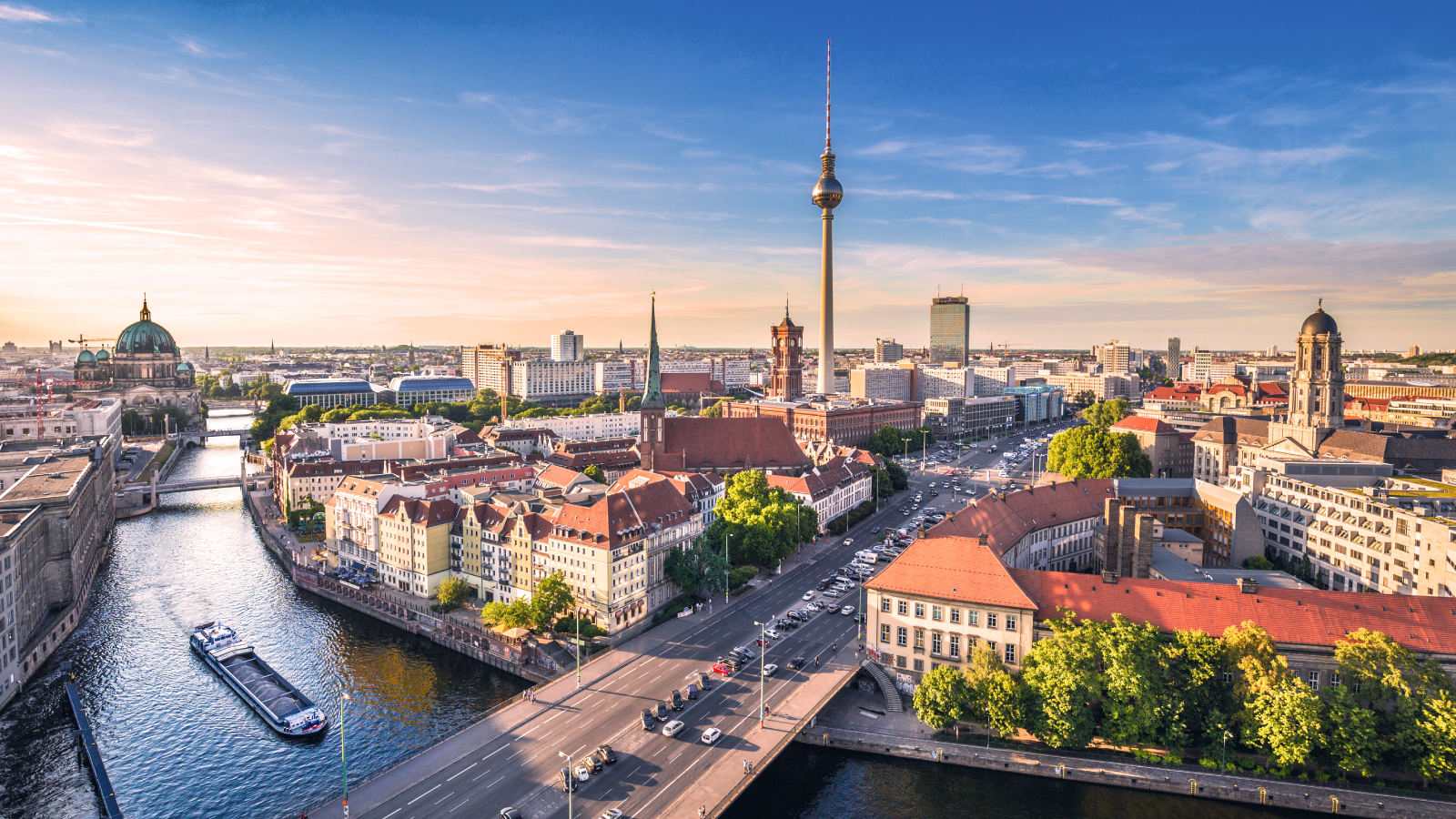 Aerial view of Berlin at sunset featuring the Spree River, Berlin Cathedral, and the iconic Fernsehturm TV tower, with historic buildings and modern cityscape under a clear blue sky.