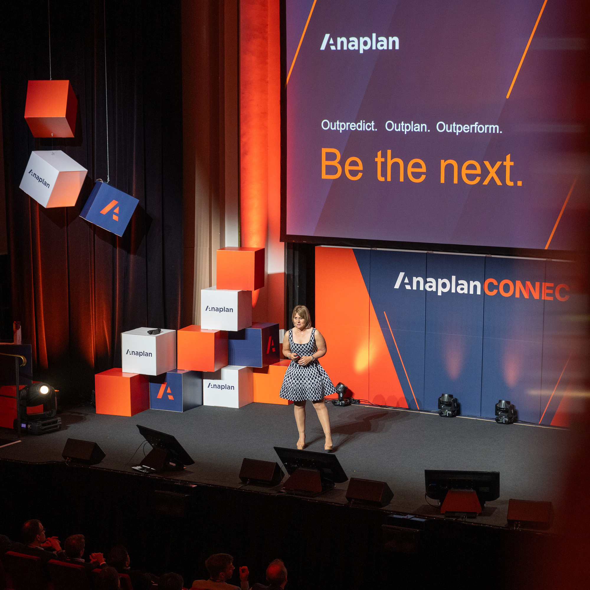 Anaplan Connect Promo Tile Anaplan Connect event photo. A speaker stands on stage in front of a large screen displaying the text “Outpredict. Outplan. Outperform. Be the next.” with Anaplan branding. The stage is decorated with stacked and hanging branded cubes in orange, navy, and white.